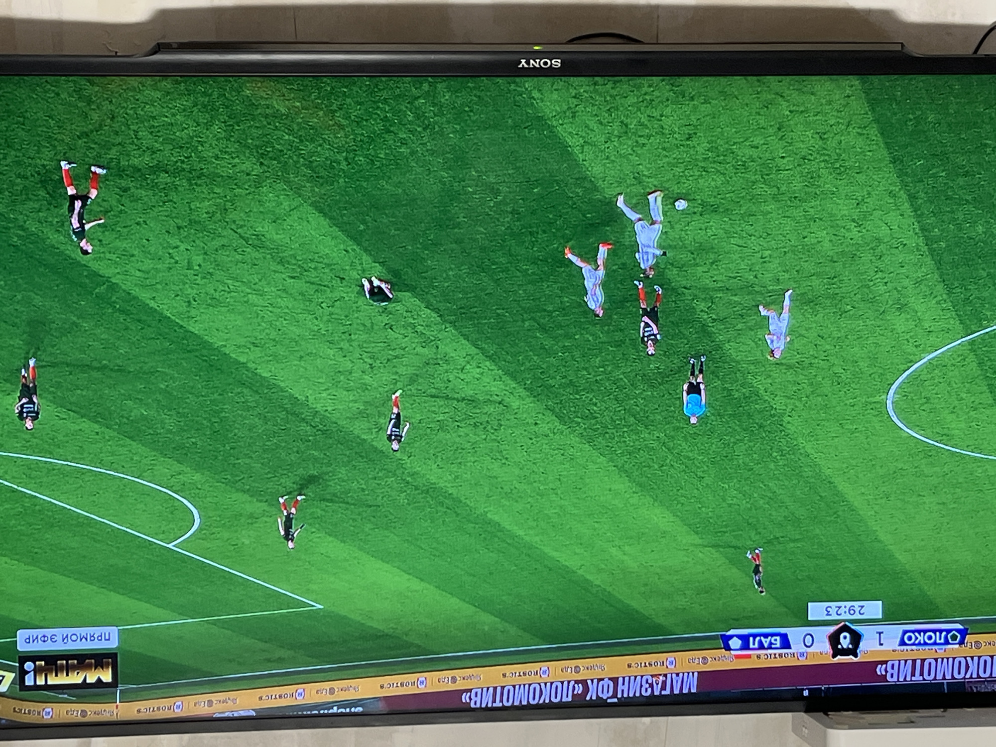The image shows a Sony television screen displaying a soccer game. The camera view is from above, showcasing a green field with players in various positions. The score is 1-0 in favor of Lokomotiv against Bal. A ticker at the bottom displays promotional messages for "Lokomotiv" and "Rostic's" (a KFC-like fast food brand). The game appears to be a live broadcast. The text in the lower-left says "live stream". The text "РОКОМОТИВ", which appears to be a misspelling of "ЛОКОМОТИВ", suggests a Russian context. The city of Chaikovsky, Russia is not explicitly visible in the image.