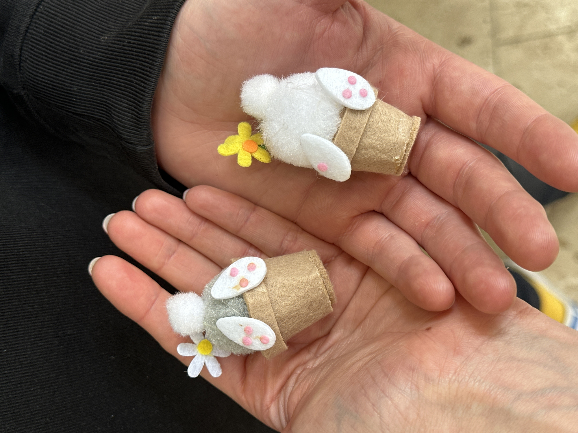 A person holds two handcrafted bunny figures in their open palms. Each bunny is made of a brown felt pot with a fuzzy white or grey bunny tail protruding from the pot. White felt feet with pink pads stick out from the sides of the pot. Each bunny has a small fabric flower attached to the side of the pot. The person wearing a black top and with manicured fingernails is in an indoor location. The city of Wealden, United Kingdom is not discernible from the image.