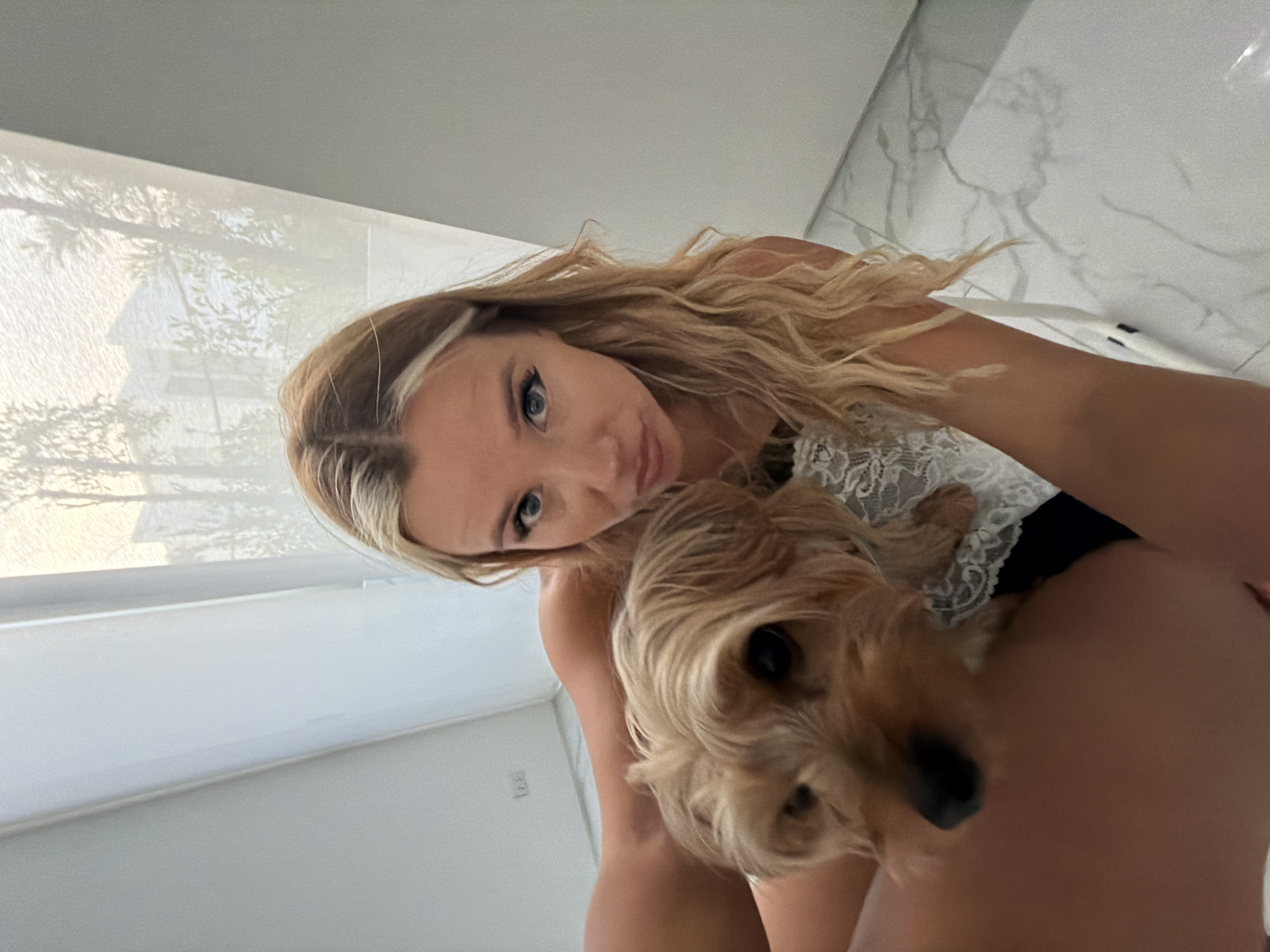 A woman with blonde hair and blue eyes is holding a small, tan dog. The woman is wearing a white lace top. The dog is facing the camera, and its face is slightly blurred. The woman is looking at the camera with a pout. The background appears to be an indoor setting with white walls and large windows with sheer curtains. The lighting suggests it is daytime. There is a tiled floor visible. The scene is likely a residential interior.