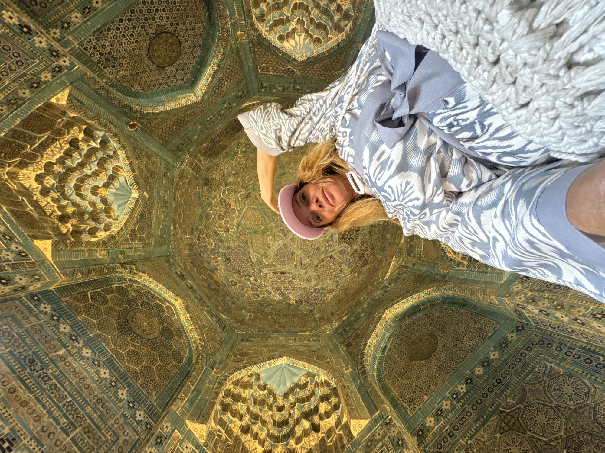 A low-angle shot shows a woman in the lower right foreground, against a highly decorated ceiling. The woman has blonde hair and wears a light pink cap. She is dressed in a patterned light grey and white wrap or robe, over a light grey garment. A white, crocheted blanket-like item is draped behind her upper body.
The ceiling is highly detailed with geometric patterns in shades of blue, turquoise, and gold. The patterns appear to be tilework and mosaics. There are archways and domes visible in the architecture.
This is a picture of the ceiling of the Gur-i-Amir Mausoleum, which is a notable building in Samarkand City, Uzbekistan.