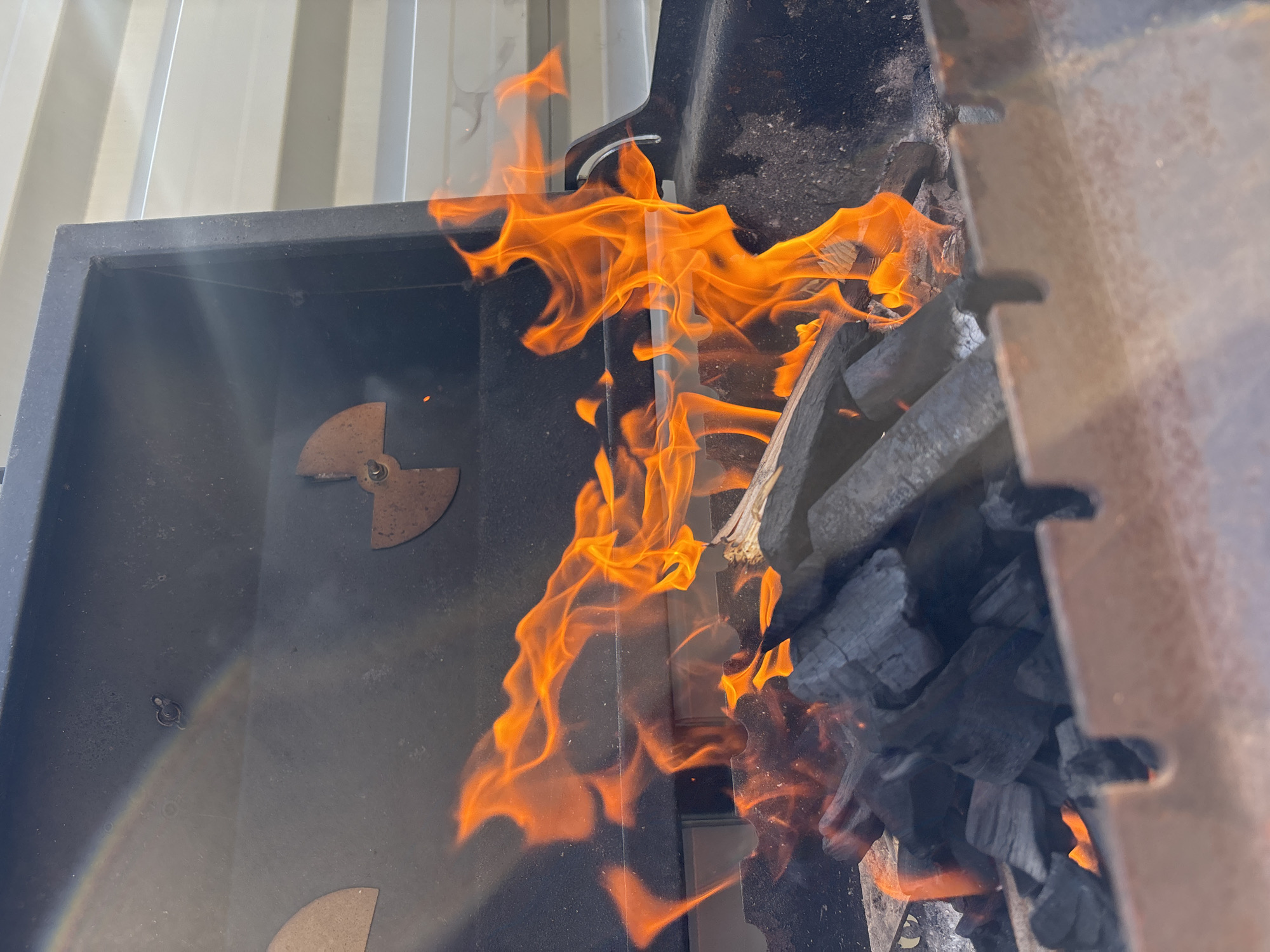 The image depicts a close-up view of a barbecue grill with active flames and charcoal. The primary subjects are the bright orange flames dancing and flickering, interspersed with dark, ashy charcoal. The grill itself appears to be made of dark metal, with some rust visible on an interior piece that resembles a damper. The background shows a pale, ribbed surface, possibly the exterior of the grill or a nearby structure, illuminated by daylight.
The activity taking place is the ignition and burning of charcoal for grilling. The vibrant flames suggest the process is underway, with heat and light emanating from the fire. Notable details include the textures of the charcoal, the fluid shapes of the flames, and the metallic sheen on the grill components. The lighting indicates it is daytime, with no specific weather conditions discernible.
There is no visible text in the image. The overall impression is one of preparation for cooking or the ongoing process of a barbecue, conveying warmth and the potential for an outdoor meal. Given the location context of Chaikovsky, Russia, this scene likely represents a common summertime activity.
