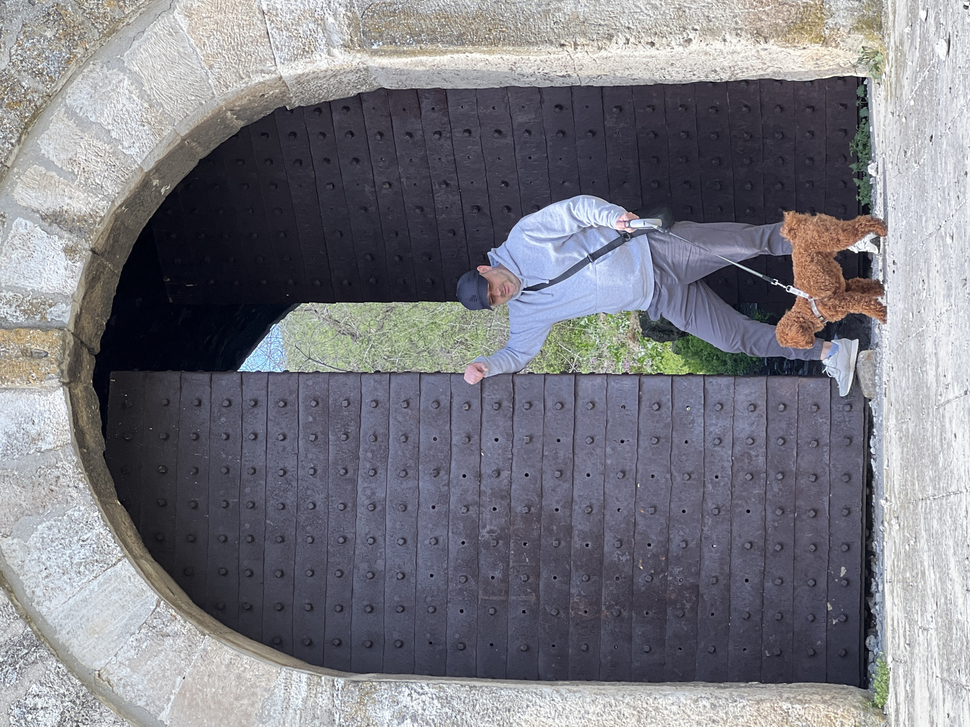 The image shows a man with a dog standing in the arched entrance of a medieval-style structure. The man is wearing a grey sweatshirt, grey pants, a black baseball cap, and light-colored sneakers. He is holding a leash connected to a small, curly-haired brown dog. They are standing in front of a heavy wooden door with metal reinforcements. The door is set into a stone archway. The background visible through the doorway suggests a natural setting with trees and possibly the sky. The image appears to have been taken in Nikopol, Bulgaria.