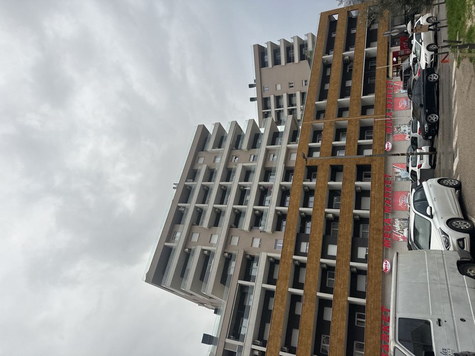 An urban scene in Budva, Montenegro, depicts a multi-story building complex under an overcast sky. The prominent structure in the foreground consists of a lower section with approximately three to four floors featuring a dark brown or reddish-brown textured facade, exhibiting horizontal rows of rectangular windows or recessed balconies, many with visible external air conditioning units. Above this, a lighter beige-colored section rises for an additional five to six floors, characterized by wrap-around balconies with glass railings. Air conditioning units are also visible on these upper residential levels. A flat roof with a clear glass perimeter railing caps the building. Behind and to the right, a second, smaller multi-story building with a similar beige facade and glass-fronted balconies is partially visible.
At street level, the ground floor of the main building displays multiple red "MEGA MARKET" signs on a white background, indicating a supermarket or large retail store. A paved parking area extends in front of the building, occupied by several vehicles. A white panel van is partially visible on the far left. To its right, a white Hyundai i20 is parked. Further right, a black BMW sedan is parked, alongside other white vehicles. A dark utility pole or street lamp is positioned in the mid-ground. On the far right, a single individual is observed walking away from the camera, holding a white umbrella, suggesting cloudy or potentially rainy weather. The sky is uniformly overcast and grey.