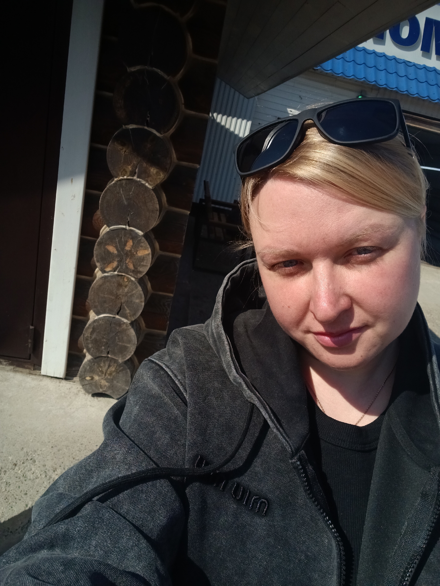 A close-up, outdoor selfie features a fair-skinned woman with blonde hair, pulled back with some strands loose, looking directly at the camera. She has dark sunglasses perched on top of her head. She is wearing a dark, faded-looking, hooded zip-up jacket over a black ribbed top, with a thin chain necklace visible around her neck. Her expression is neutral with a subtle, slight smile.
The setting is outdoors on a sunny day, indicated by bright natural light creating clear shadows. The background to the left shows a section of a building constructed from horizontal wooden logs, characteristic of a log cabin or traditional Russian architecture, alongside a white vertical beam or door frame. To the right, above the woman's head, part of a blue corrugated metal roof or sign is visible, featuring white letters that spell "OM", possibly part of a larger word or brand. The ground visible in the bottom left is concrete or paved.
On the woman's jacket, near the zipper on the left side, there is an embossed or stitched word that appears to be "turum". The scene captures a casual moment in a setting that blends rustic architectural elements with potentially commercial or public spaces, fitting the context of Chaikovsky, Russia.