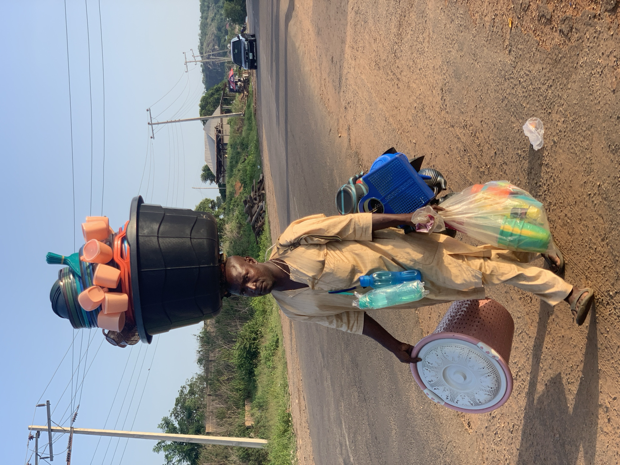 A man stands on a paved road in Gbeleko, Nigeria, carrying a large assortment of household items. He has a black bucket balanced on his head, filled with peach-colored cups and various other plastic containers. In one hand, he holds a plastic bag containing colorful plasticware, and a blue water bottle is slung over his shoulder. In his other hand, he carries a round, lacy-patterned laundry basket. The man wears a light brown outfit and sandals. The road runs through a rural area, with vegetation visible on either side and power lines stretching overhead. In the background, there are houses and a vehicle on the road.