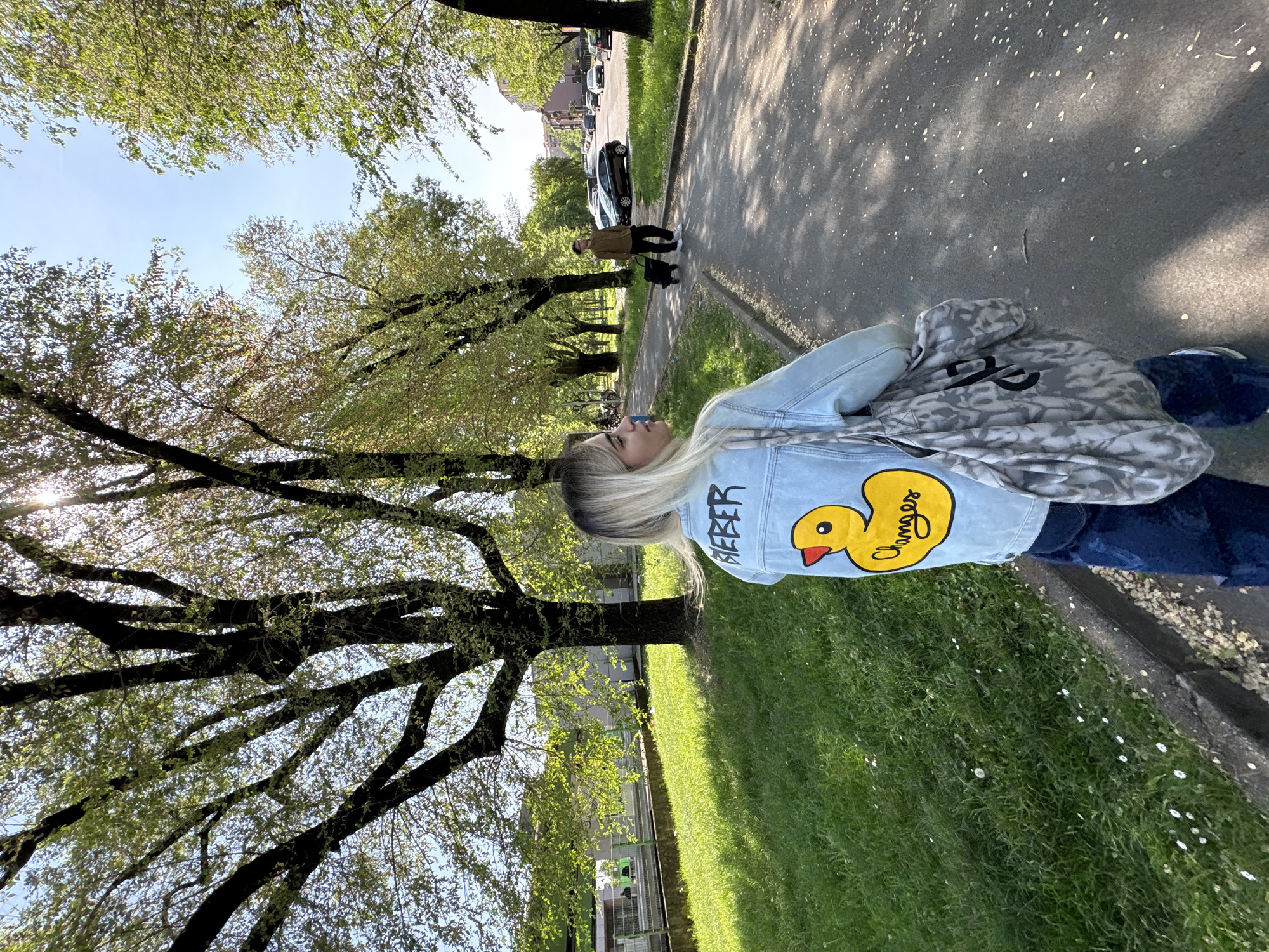 The image depicts a person walking along a sidewalk in what appears to be a park-like setting. The person is wearing a light blue denim jacket with a cartoon duck design and the word "Bieber" on the back, paired with dark blue jeans. A patterned grey and black bag is slung over their shoulder. The person has blonde hair and is facing towards the left of the frame.
To the right of the person is a strip of grass, with small white flowers scattered throughout. To the left is a line of mature trees with dense foliage, casting shadows on the sidewalk. Further down the sidewalk, two individuals, one with a dog, can be seen walking in the same direction. Cars are parked on the street beyond them.
Based on the style of architecture and urban landscape, the image location is the city of Milan, Italy.