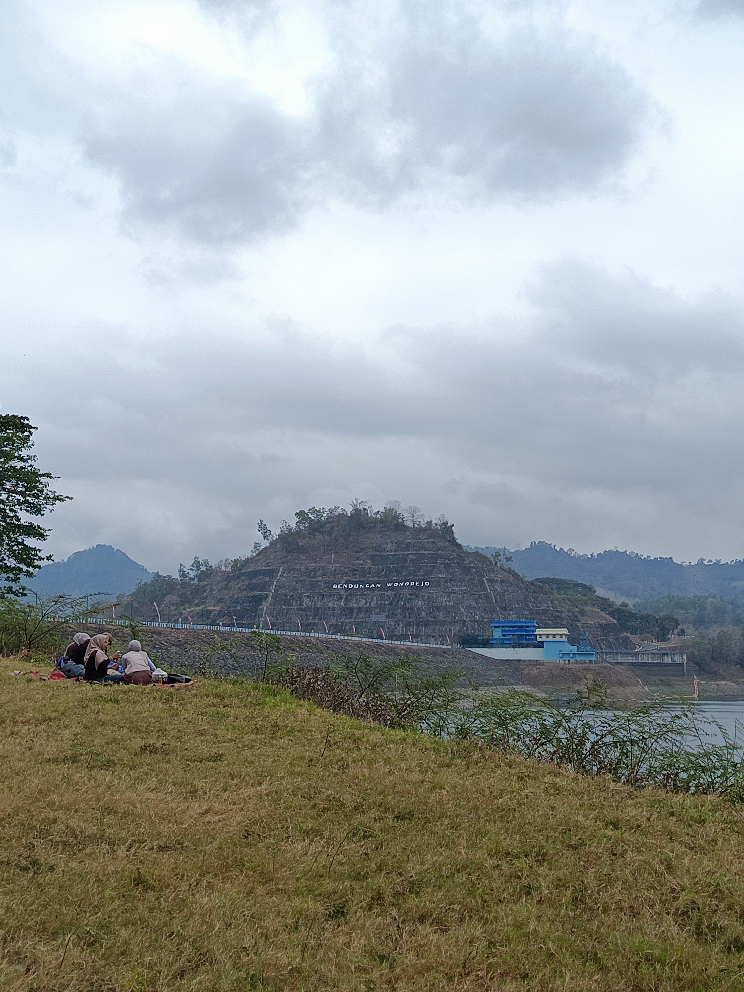 Three individuals, appearing to be women wearing hijabs, are seated on red blankets on a grassy hillside, facing away from the viewer and towards a large dam. The dam is carved with the text "BENDUNGAN WONOREJO" and has a blue and white building complex at its base. Beyond the dam, a body of water is visible, with hills and mountains forming the backdrop under a cloudy sky. The foreground consists of dry, yellowish grass and scattered green bushes. The overall scene depicts a quiet outdoor gathering at a scenic location in Segawe, Indonesia.