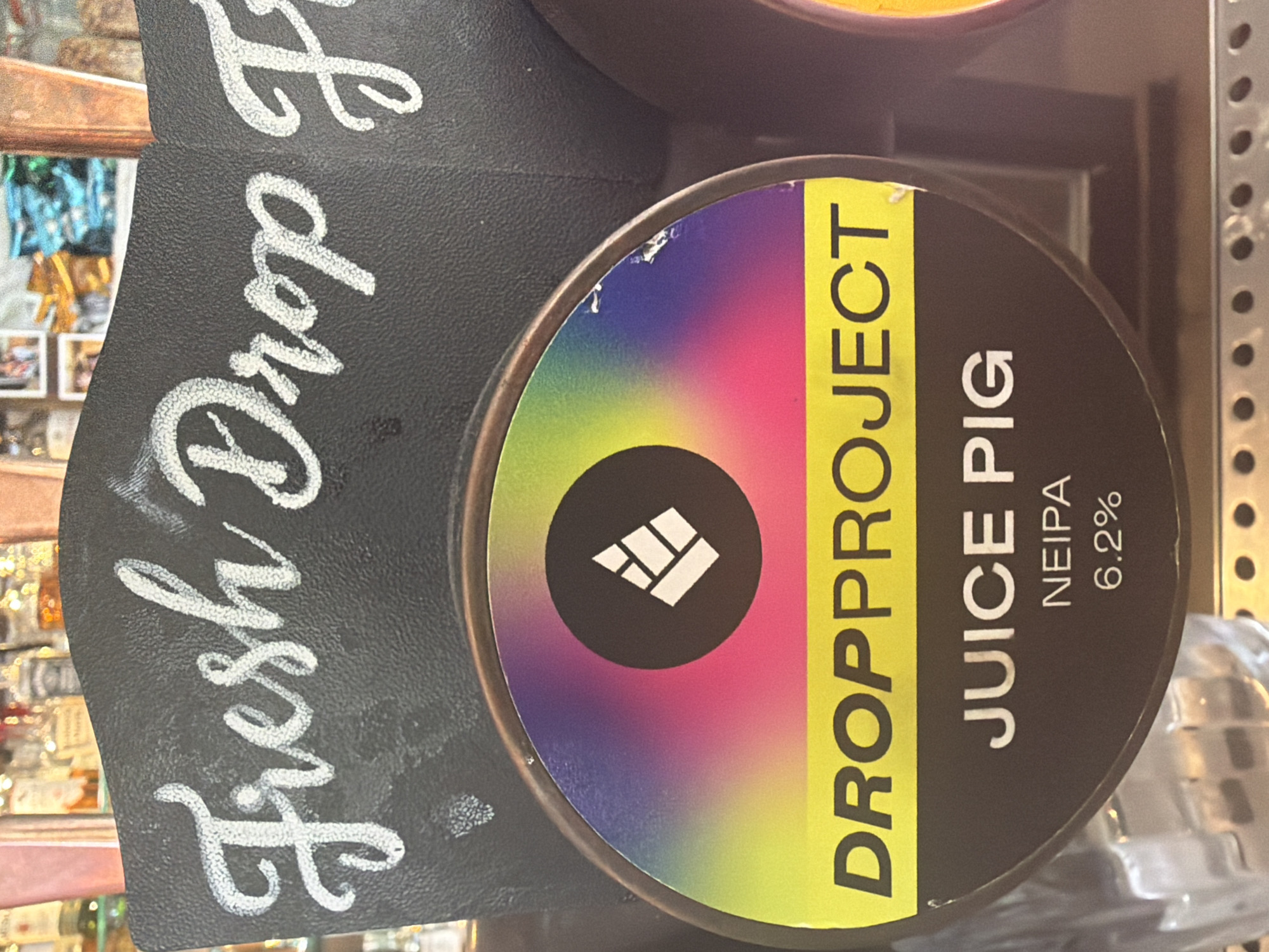 The image shows a close-up of a beer tap at a bar, likely in Epsom and Ewell, United Kingdom. The tap has a circular label with a colorful gradient and a white logo. Below the logo, the words "DROPPPROJECT" are written in yellow text on a yellow band. Further down, the text "JUICE PIG" is written in white. Below that, "NEIPA 6.2%" indicates the type of beer and its alcohol content. The background shows a chalkboard with white handwritten text that appears to say "Fresh Drop P". Behind the tap, blurred bottles of alcohol and bar equipment are visible.