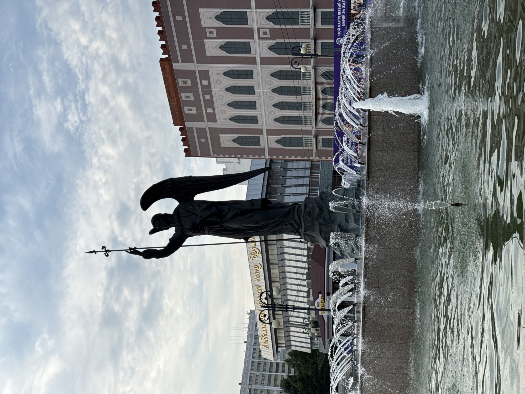 The image depicts a statue of an angel holding a spear, positioned in front of a fountain in Yoshkar-Ola, Russia. The statue is dark in color and appears to be made of metal. The fountain has multiple jets of water spraying upwards, creating a misty effect. In the background, there are several buildings, including a theater with ornate architecture and a multi-story residential building. The sky is cloudy, with patches of blue visible. The overall scene suggests an urban public space, possibly a plaza or park.