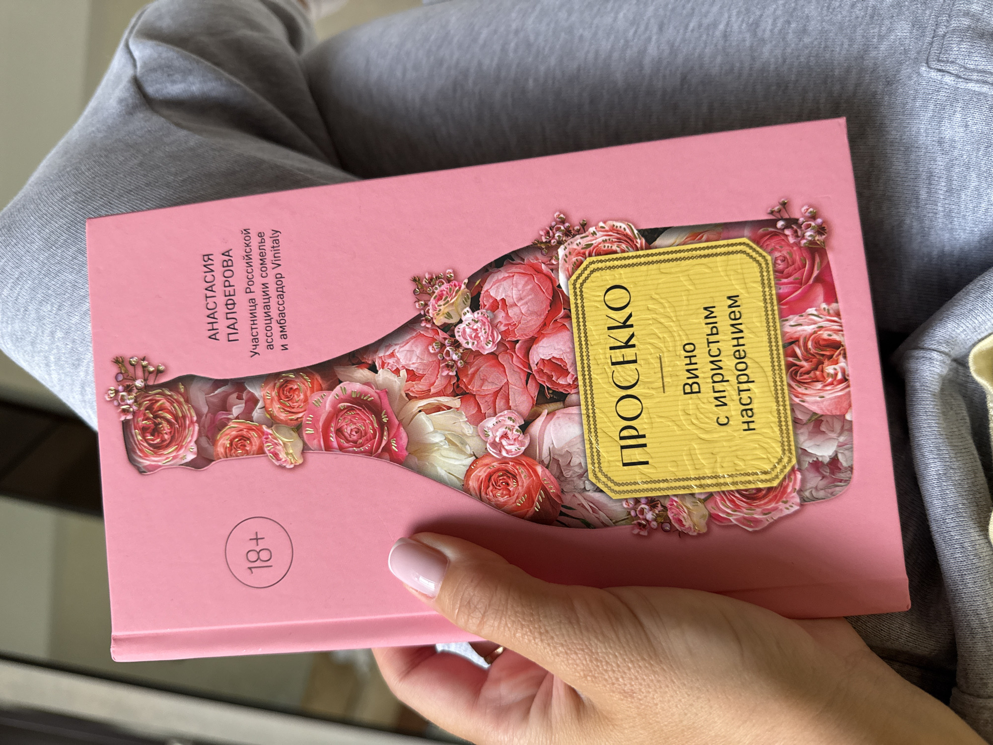 A person in gray sweatpants is holding a pink book titled "Prosecco" in Russian. The book has a cutout in the shape of a bottle filled with floral imagery. The text on the book cover reads "Anastasia Palferova, Participant of the Russian Sommelier Association and Ambassador Vinitaly". There is an "18+" symbol in a circle. The image was likely taken indoors. There is no indication of the city of Sochi in the image.