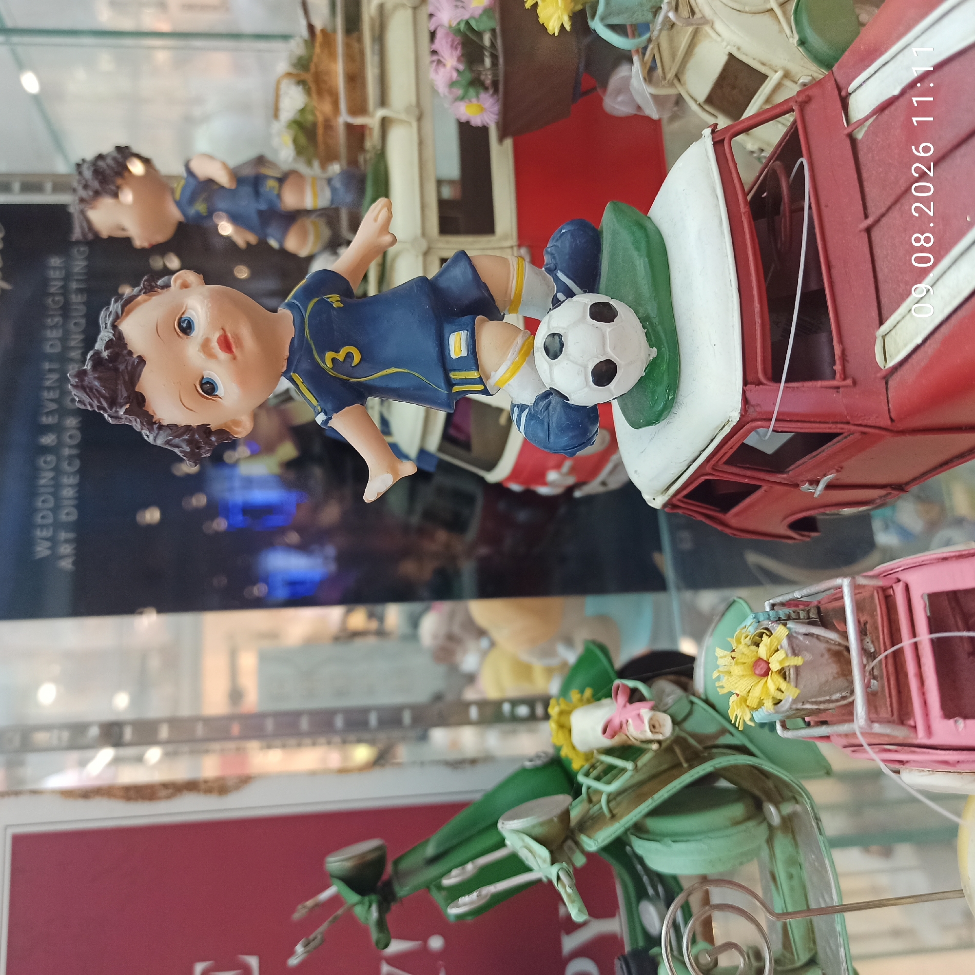 A close-up, eye-level shot captures a display of miniature decorative items, possibly within a shop window or a curated exhibit, featuring a predominantly red and green color palette. In the foreground, a red toy car, resembling a classic station wagon, is prominent. On top of this car rests a figurine of a young boy dressed in a blue soccer uniform with the number "3" on his jersey, appearing to be mid-kick on a green surface with a white soccer ball. To the left of the red car, a mint green, vintage-style scooter is parked, adorned with a small bouquet of yellow flowers and a pink ribbon tied to its handlebars. Behind these primary subjects, other decorative items are visible, including baskets with flowers and another figurine of a boy in a similar soccer uniform, slightly out of focus. The background is blurred, showing reflections on a glass surface, with faint text suggesting "WEDDING & EVENT DESIGNER ART DIRECTOR BANQUETING." A timestamp, "09.08.2026 11:11," is superimposed on the lower right corner of the image. The scene is static and does not depict any ongoing actions or interactions. The location in Molfetta, Italy, is not discernible from the visual information provided.