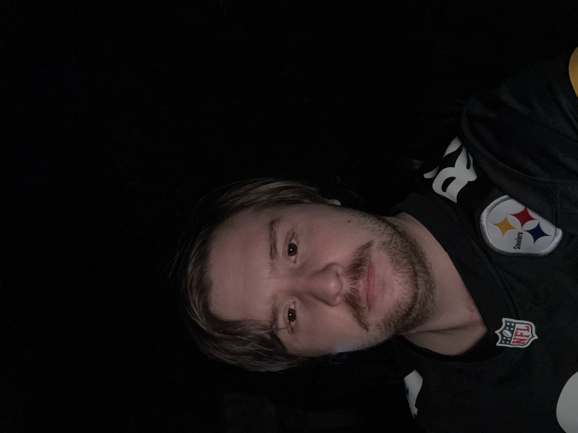 The image is a close-up, front-facing selfie of a man in a Pittsburgh Steelers jersey. He has light brown hair, a mustache, and a light beard. The background is dark, suggesting it was taken at night or in a dimly lit environment. The man is wearing a black jersey with the NFL logo on the left chest and the Steelers logo on the right. The lighting appears to be coming from the front, illuminating his face. No specific location in Cañada de Madero, Mexico, can be identified from the image.