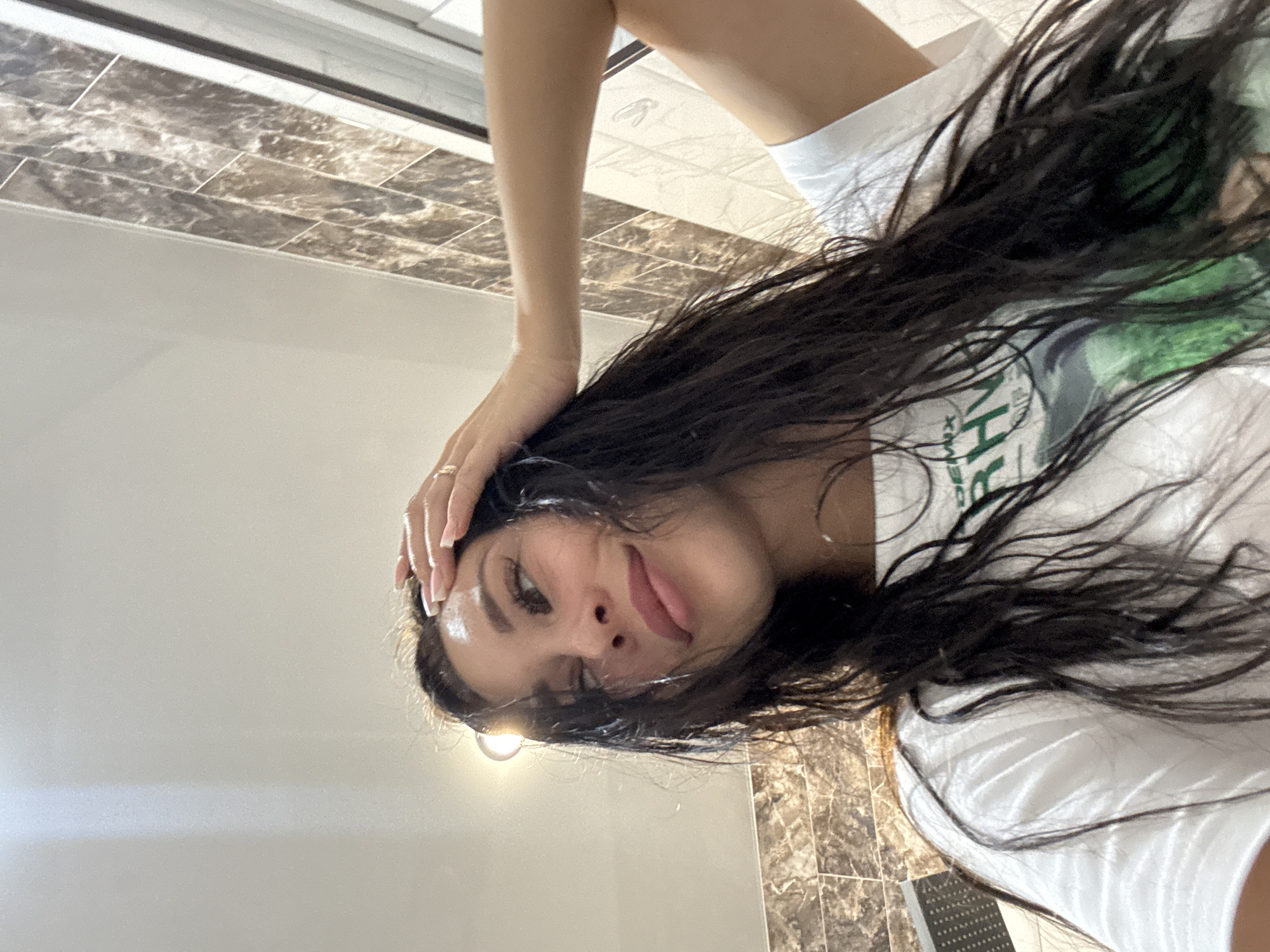 A woman with dark hair wearing a white t-shirt is posing for a selfie indoors. The background shows a bathroom with brown tiled walls and a white bathtub. The location is likely in Russia, but the city of Chaikovsky cannot be confirmed from the image.