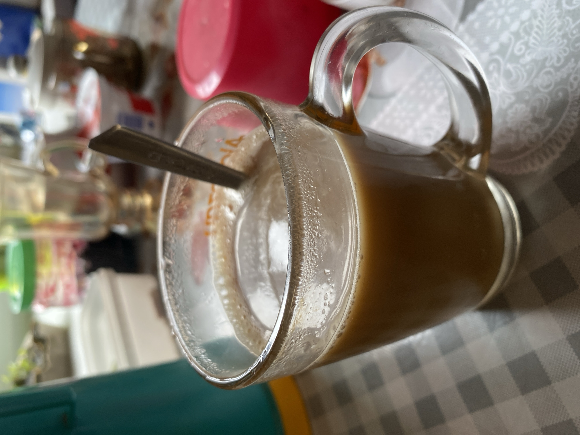 A close-up shot depicts a partially consumed beverage in a clear glass mug, with a spoon inside, placed on a tablecloth with a grey and white checkered pattern. The beverage appears to be coffee with milk or creamer, showing condensation on the mug's surface. In the blurred background, other objects are visible, including what seems to be cups and possibly other kitchen items. The location appears to be an indoor space.