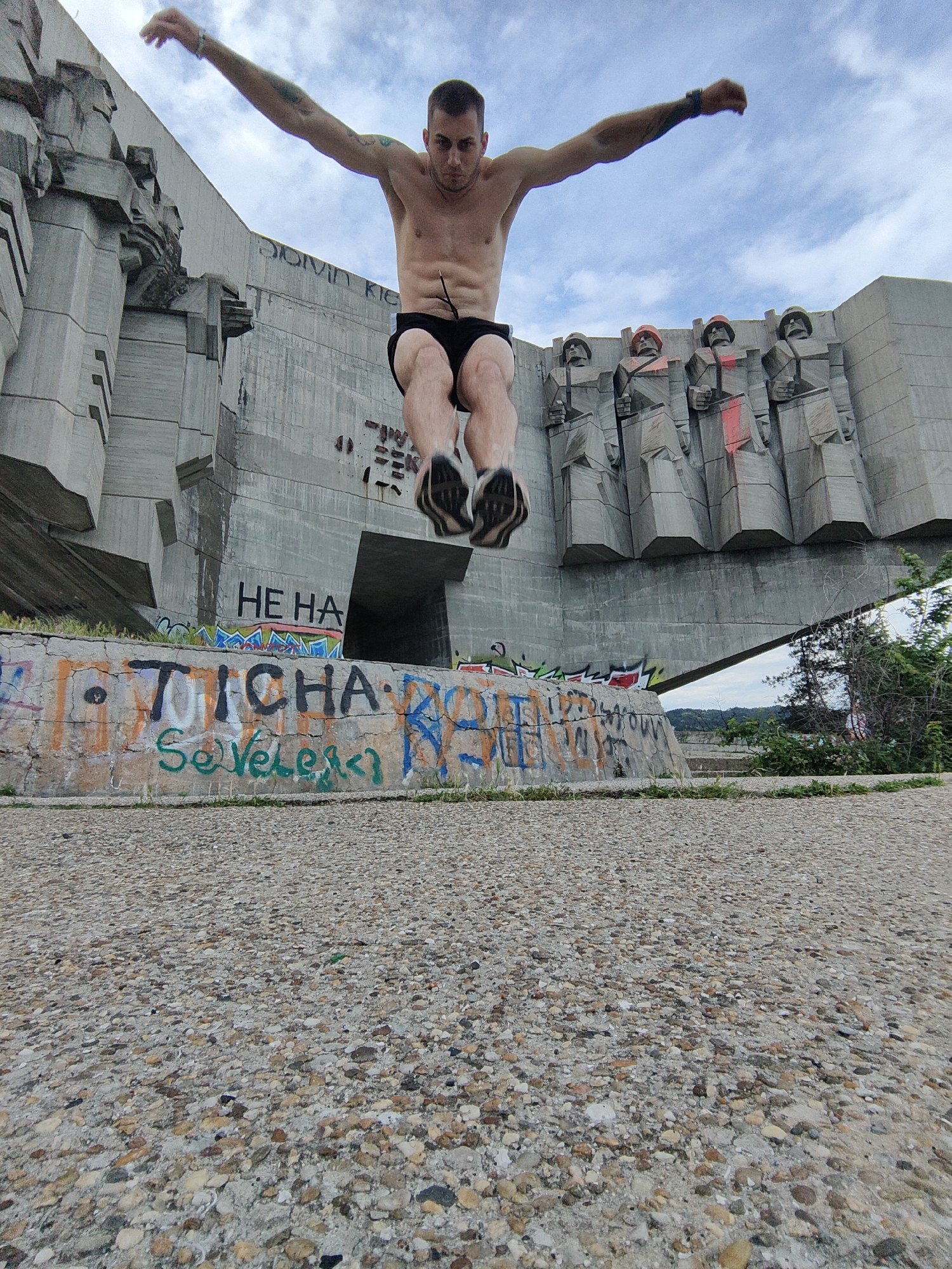 The image features a muscular, shirtless man in mid-air, captured during a jump or parkour movement. He wears black shorts and black-and-white sneakers, with his arms outstretched and legs bent, showcasing tattoos on his arms. His face is concentrated as he performs the action.
The setting is the Monument of Bulgarian-Soviet Friendship in Varna, Bulgaria. Behind the man, the imposing, brutalist concrete monument dominates the background. The structure features abstract, angular shapes on its left side and a row of sculpted figures, likely soldiers, on the right. These figures are depicted with helmets, and several have noticeable red paint marks on their chests and headwear, indicating vandalism.
Below the main monument structure, a concrete retaining wall is heavily covered in colorful graffiti. Visible text includes "HE HA" on a cleaner part of the monument's lower wall. On the graffiti-covered wall, distinct words like "TICHA" and "SeVeles<3" are discernible among numerous other tags and scribbles. Faintly visible higher on the monument's wall are the letters "CALVIN KLE," likely part of "CALVIN KLEIN."
The foreground consists of a rough, pebbled concrete surface. The sky above is overcast with scattered clouds, suggesting a daytime scene. Beyond the monument, glimpses of green foliage and a distant, hazy landscape can be seen. The image blends historical architecture with contemporary urban elements and human athleticism.