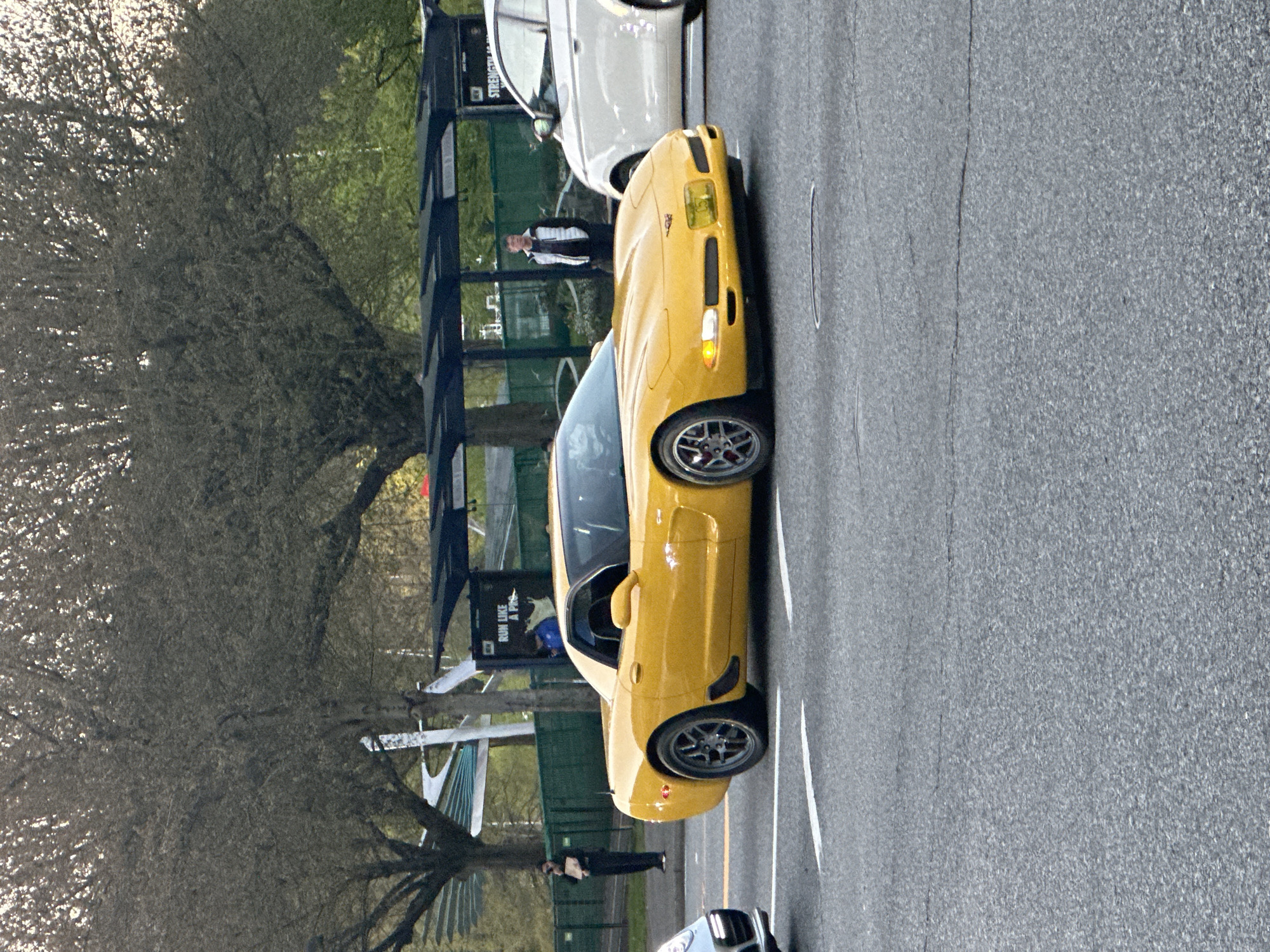 A bright yellow Chevrolet Corvette C5 sports car is the central subject, positioned in the midground and angled slightly left. Its front turn signal is illuminated, and its headlight covers are distinctly yellow-tinted. The car features aftermarket or performance-oriented wheels.
The scene unfolds on an asphalt road with visible white painted lines in the foreground and midground. In the background, several people are present: one individual stands near the front of the yellow Corvette, and another is visible further back near a dark, modern-looking structure. More people are partially obscured behind this structure. A white car is parked directly behind the Corvette, and a portion of a dark vehicle is visible in the bottom right corner of the frame.
To the right side of the image, large deciduous trees with sparse, budding foliage are prominent, suggesting spring or early summer. The dark structure in the background, possibly a bus shelter or an event booth, features visible text.
The overall setting appears to be a public outdoor area, potentially a street or a gathering point for a car event. The lighting is diffused, indicating an overcast day or perhaps early morning/late afternoon.
Visible text on the dark structure reads "RUN LIKE A PRO," with the word "STREET" partially visible above it.