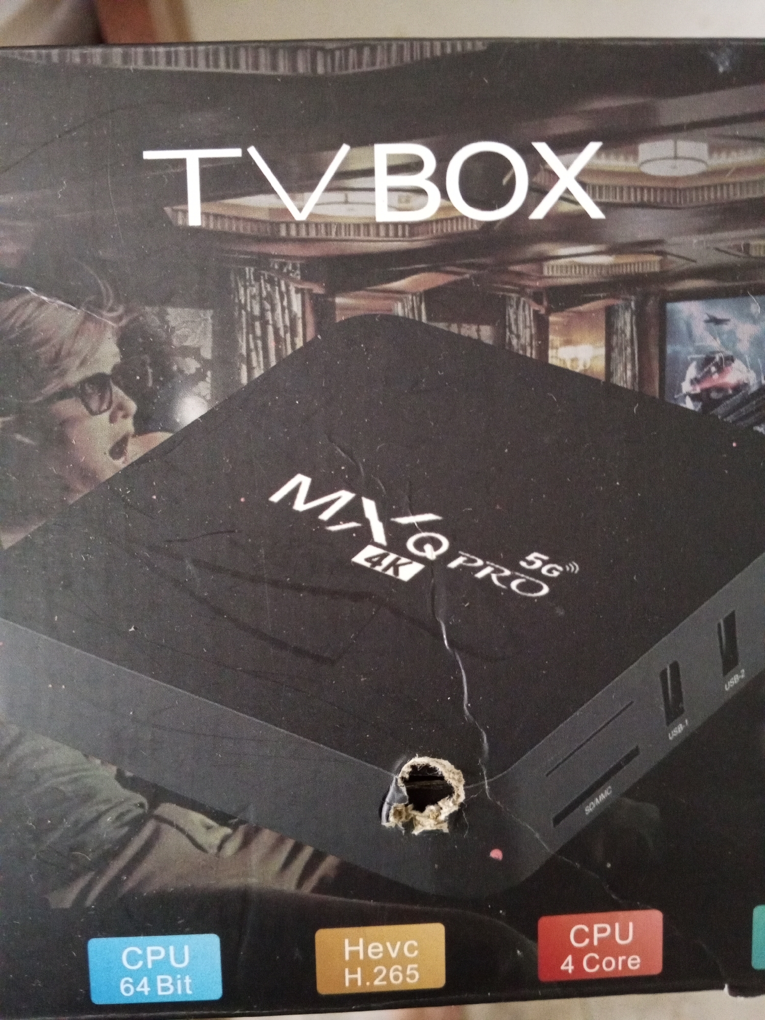 The image shows a damaged box for an MXQ Pro 4K TV box. The box is black with the words "TVBOX" in white at the top. Below, the brand name "MXQ Pro" and "4K" are printed, along with "5G". The box has a small hole on the left side. At the bottom are color-coded labels indicating the features: "CPU 64 Bit," "Hevc H.265," and "CPU 4 Core." The background behind the box shows an image of a person wearing glasses and an interior with architectural details and screens showing a space scene. The image appears to be taken indoors. No location information is available.