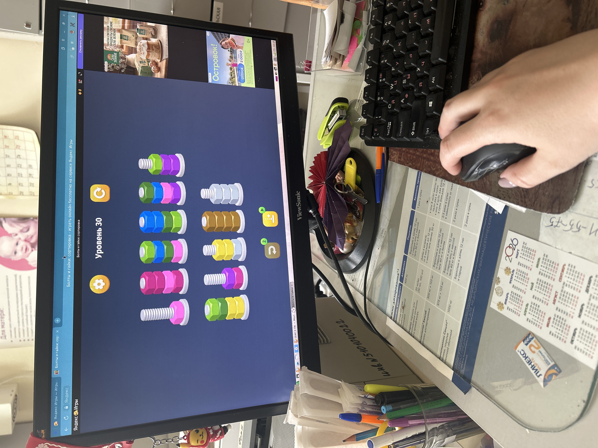 The image captures a cluttered workspace with a focus on a computer monitor displaying a puzzle game called "Bolts and Nuts: Sorting" on the Yandex Games platform. The screen also shows advertisements for "Easter" products and "Ostrovok! Vacation" in Russian.
A person's hand is visible, using a computer mouse on a patterned mousepad next to a keyboard with Cyrillic letters. The desk is covered with various items, including pens, a stapler, keys, and documents, suggesting a busy office environment. A small calendar for March 2026, featuring the branding "ЛИНЕКС ФОРТЕ 2," is also visible. Additionally, there's a document with medical instructions written in Russian.
While the language used suggests Russia, there is no concrete information that indicates the precise location as Фоки. The environment is indicative of a typical office setting, albeit somewhat disorganized. The scene is captured from a close perspective, with a concentration on the computer screen and immediate workspace.
