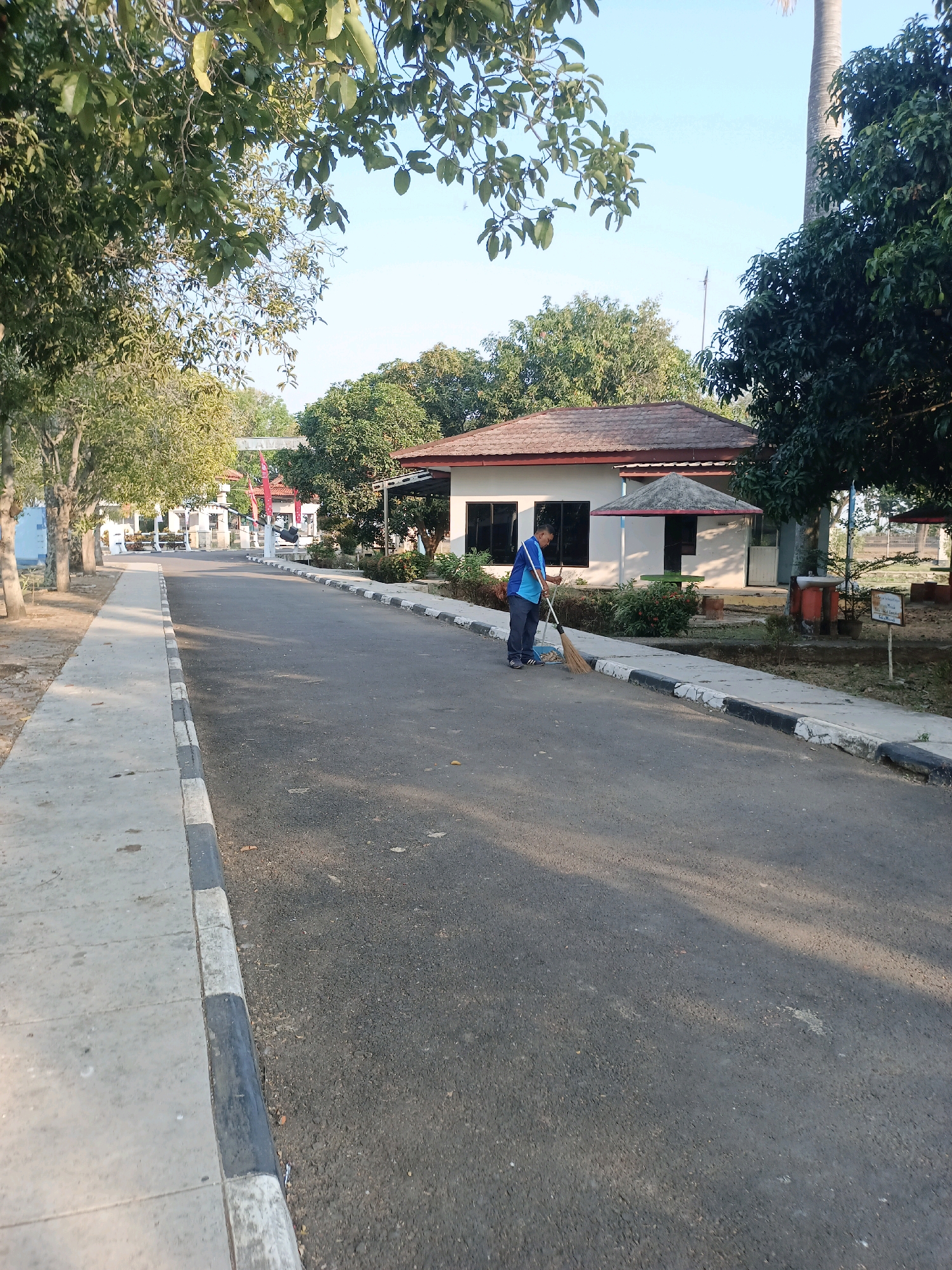 A lone man is sweeping a paved road in front of a small building in Subang, Indonesia. He is wearing a blue shirt and grey pants and is holding a broom. The building has a brown roof and white walls. There are trees and a sidewalk on the left side of the road. The sky is clear and blue. The overall scene depicts a clean and orderly environment.