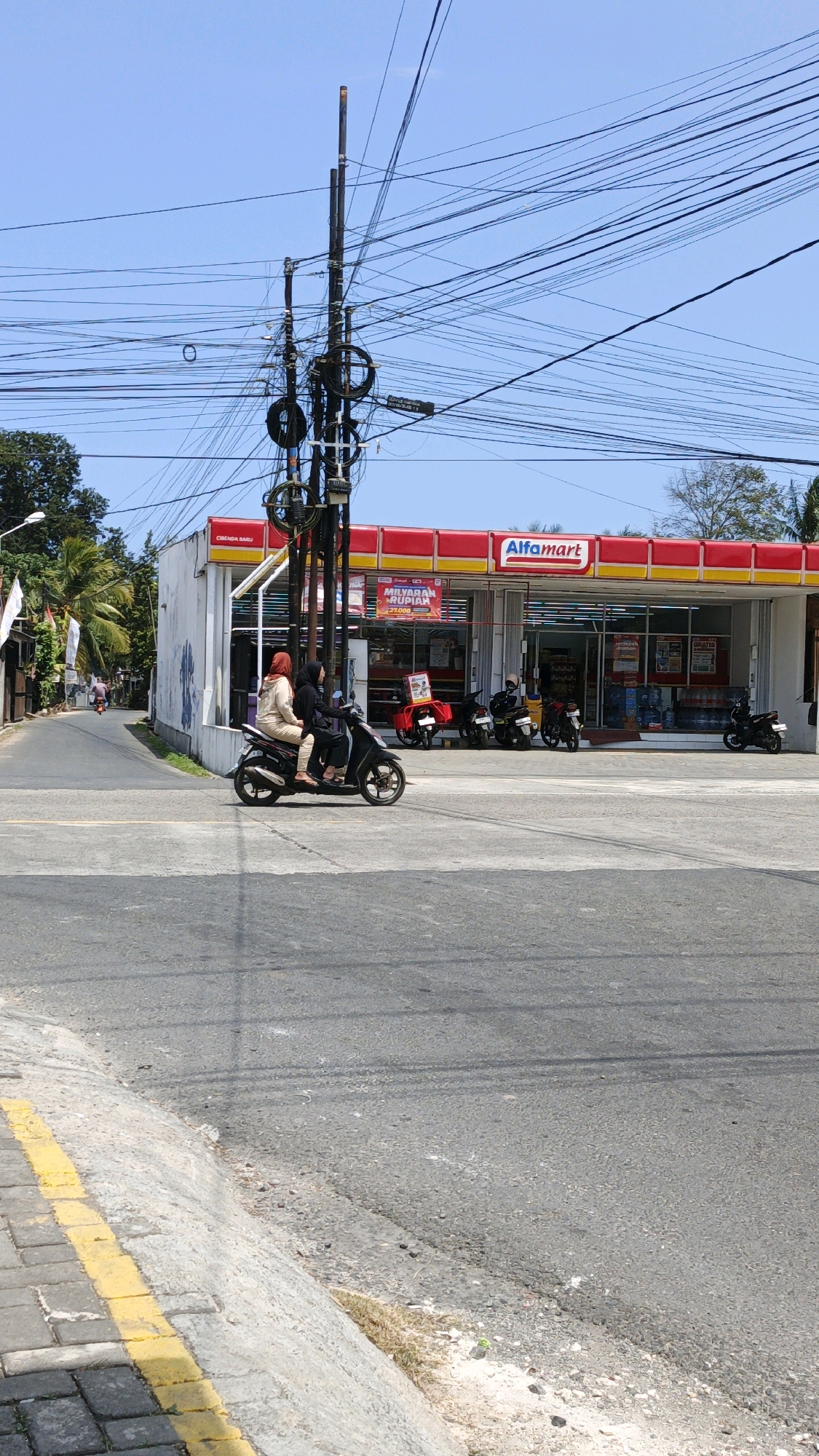 A sunny day scene in Cibenda, Indonesia, captured on a street with a bright blue sky. A prominent Alfamart store with a red and yellow awning is visible in the background, featuring various products displayed inside and promotional posters outside. In front of the store, several motorcycles are parked.
In the foreground, two women are riding a black motorcycle. The woman in the back is wearing a black hijab and a dark top, while the woman in the front is wearing a light-colored hijab and a beige outfit. They are traveling on a paved road that transitions to a sidewalk with yellow markings.
The street is lined with utility poles and an intricate network of overhead wires, indicative of an urban setting. Trees and some buildings are also visible along the street, contributing to the streetscape. The overall image portrays a typical street scene in the area.