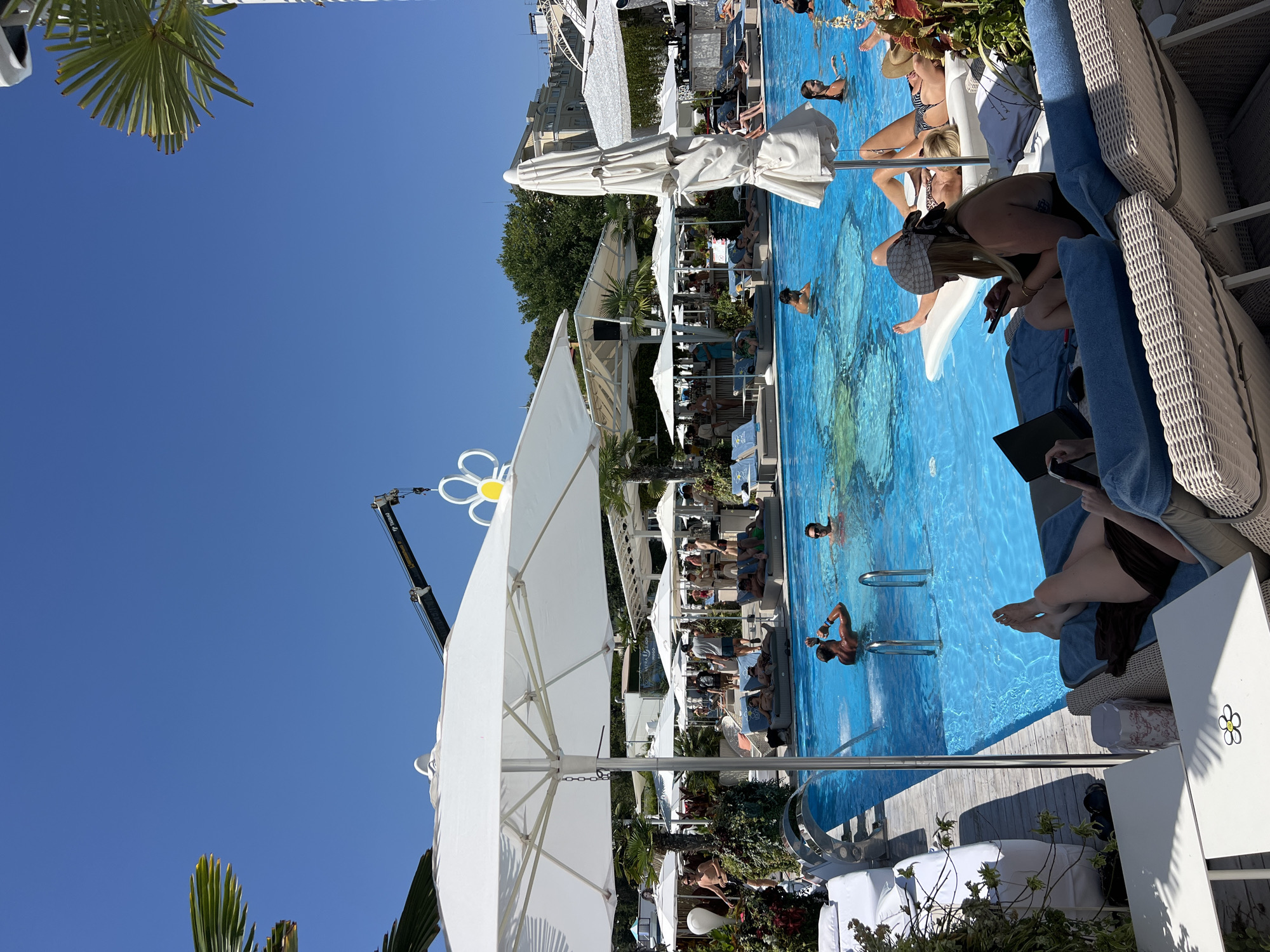 The image depicts a sunny day at a crowded outdoor swimming pool complex, likely a beach club or resort, in Odesa, Ukraine. The pool itself is filled with bright blue water, and numerous people are visible swimming, lounging on floaties, or standing in the water. Several white umbrellas and loungers surround the pool, providing shade and seating. In the background, there are more people, cabanas, palm trees, and buildings, suggesting a vibrant recreational area. A construction crane is also visible in the sky above the complex. The overall atmosphere appears to be one of leisure and entertainment under a clear blue sky.