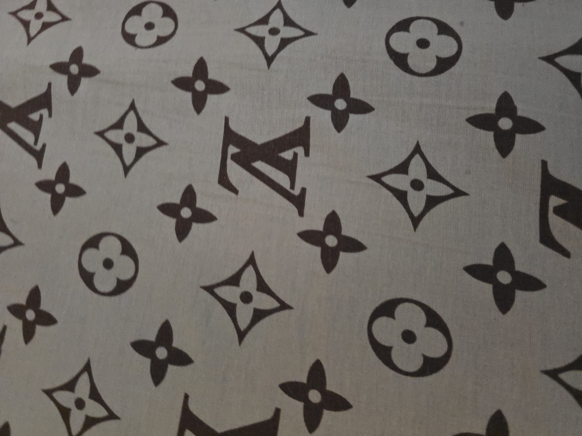 A close-up, top-down shot shows a light gray fabric with a repeating pattern of dark brown Louis Vuitton monogram symbols. The pattern includes the LV initials, four-petal flowers, and circular flower-like shapes. The fabric appears to be a soft material like silk or cotton, and there are subtle wrinkles and folds visible, indicating it might be draped or a part of a garment. The lighting is even, suggesting an indoor environment. The image is focused on the pattern itself, with no other objects or subjects present. The location is unknown.