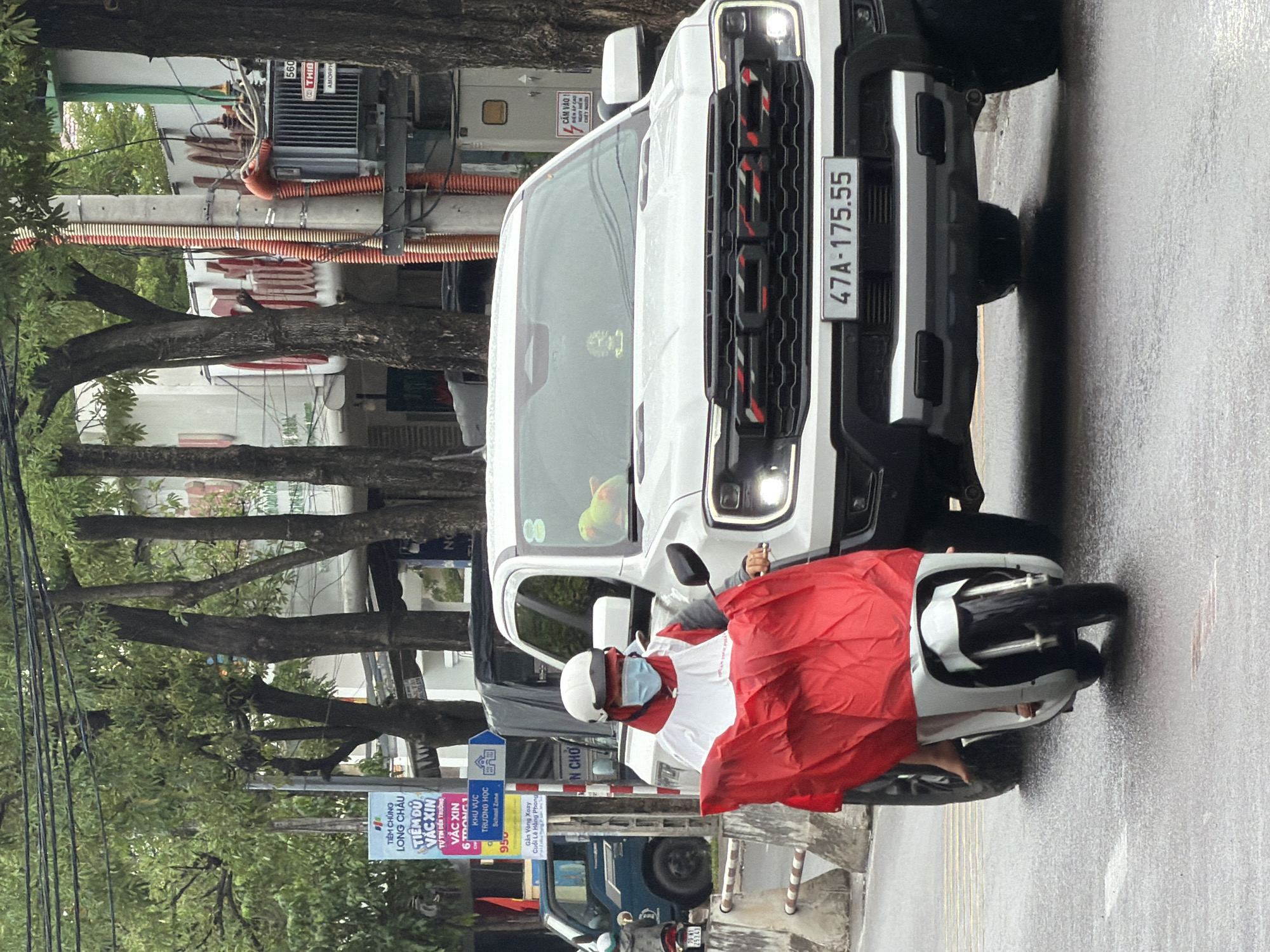 The image depicts a street scene in Nha Trang, Vietnam, with a white Ford Ranger Raptor pickup truck stopped in the foreground. A person on a white electric scooter is in the process of passing the truck on the left. The scooter rider is wearing a white helmet, a blue face mask, and a red and white jacket, and is holding a large red fabric over their lap, obscuring their lower body. The scooter has a white front fairing and black wheels. The truck has a license plate reading "47A-175.55" and a distinctive black grille with red and white accents. A plush toy parrot is visible on the dashboard of the truck. The street is wet, indicating recent rain, and there are trees lining the road. In the background, buildings and utility poles are visible. A blue truck is parked on the left side of the image, behind the scooter. A billboard with Vietnamese text is also visible on the left.