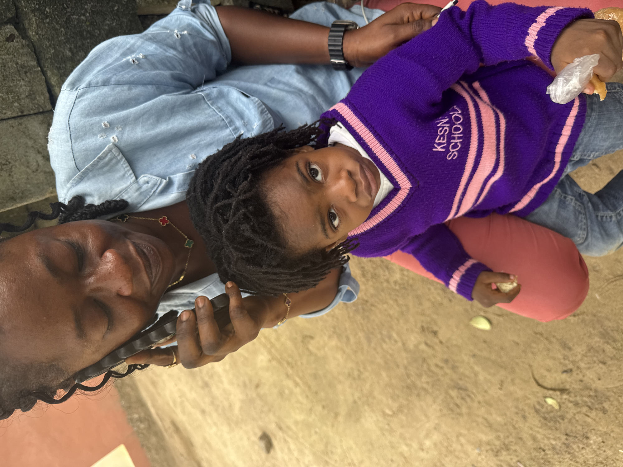 In Ganyingbo, Nigeria, a Black woman is on a phone call, holding a dark-colored smartphone to her left ear with her right hand. She is wearing a light blue denim shirt with distressed details and a gold necklace with clover pendants. To her right, a young Black child sits on her lap, wearing a purple V-neck sweater with pink stripes and the text "KESN SCHOOL" embroidered on the left chest. The child is looking directly at the camera with a serious expression and holds a piece of bread wrapped in plastic in their right hand. The child is also wearing blue jeans. The background consists of a textured, sandy ground and a muted orange wall. The scene is captured from a high-angle perspective, looking down on the subjects.