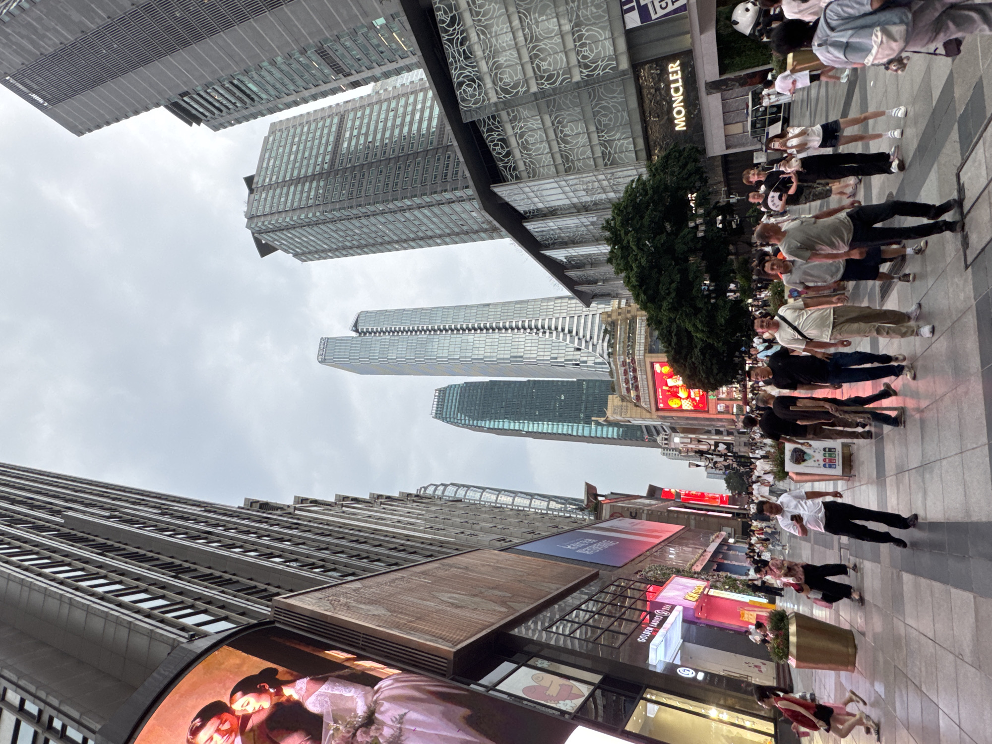 The image depicts a street scene in the Yuzhong District of China, characterized by towering modern architecture and a bustling pedestrian thoroughfare. The perspective is low, looking up at the imposing skyscrapers that flank a wide, tiled walkway. On the left, a large digital billboard displays an image of a couple, likely promoting a fashion brand, with the text "GOLDEN LADIES" visible on a storefront below. The buildings are sleek and contemporary, featuring extensive glass facades and geometric designs. Several tall, slender skyscrapers dominate the background, some with distinctive architectural elements like curved glass or patterned exteriors.
The walkway is populated by numerous pedestrians of various ages, dressed in casual summer attire. People are walking in different directions, some individually, others in small groups. Many are looking at their phones, while others are engaged in conversation or simply navigating the crowded space. There are large decorative planters along the sides of the walkway, one of which is a prominent metallic gold cylinder. A white vehicle, possibly a delivery van or service car, is visible parked on the right side of the street, near a building with the prominent "MONCLER" logo displayed on its facade. A large decorative image of a panda is also visible near the Moncler store. The sky is overcast, suggesting a cloudy day. The overall impression is of a vibrant and active urban commercial district.