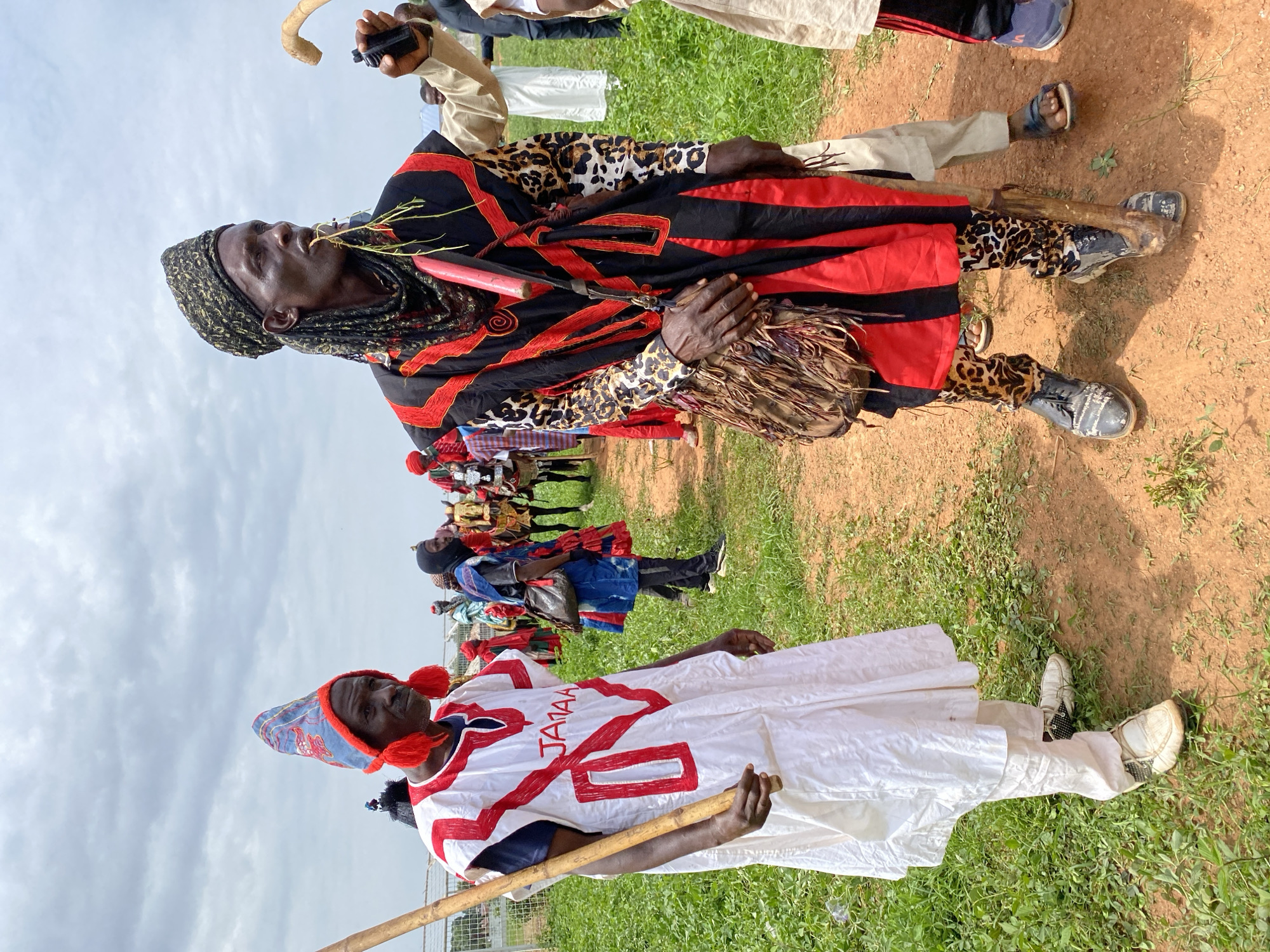 In Kafanchan A, Nigeria, two men stand out in a crowd on a field of green grass bordered by a patch of red dirt. The sky is overcast.
One man wears a predominantly white robe decorated with red patterns and the word "JAMAA" written on it. He holds a wooden staff. The other man wears a black and red patterned shawl over a leopard-print top and pants. He holds a bundle of twigs near his mouth and carries a mask.
A crowd of similarly dressed individuals can be seen in the background.