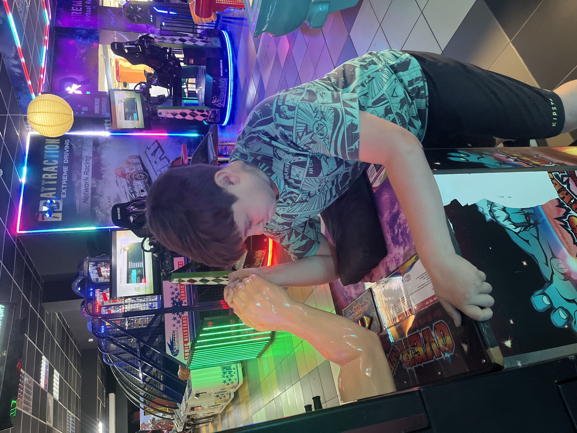 An upright, eye-level shot captures a young person immersed in an arcade game within a dimly lit entertainment center, likely in Krasnodar, Russia, given some of the Cyrillic text visible. The central subject, a young person with short brown hair, wears a blue/teal t-shirt with comic book-style patterns and black "KIPSTA" brand shorts. They are leaning over an arcade machine, their hands positioned near a prominent, large, beige, muscular arm with a clenched fist, suggesting an arm-wrestling or strength-testing game titled "OVER THE TOP." Their expression shows focus and concentration.
The background is a vibrant tapestry of neon lights in blue, pink, and green, emanating from various arcade cabinets. A yellow spherical lantern hangs in the upper left, adding a warm glow. Other games are visible, including racing simulators with text like "ATTRACTION," "EXTREME DRIVING," "Network Racing," "BRIDE," and "MCRAE," along with Russian text "РАЛЛИ-КРОСС" (RALLY CROSS). Further back, signs for "Virtual Reality" and "IMMERSE YOURSELF" are seen. A "SONIC ALL-STARS" basketball game is also partially visible. The floor features a black and white checkered pattern in some areas. The scene depicts an energetic, fun-filled atmosphere typical of an arcade, likely taking place in the evening or night. Other legible text includes "RAPER," "REAP," "ANTE," "DO/360," and "www.xdot.ru."