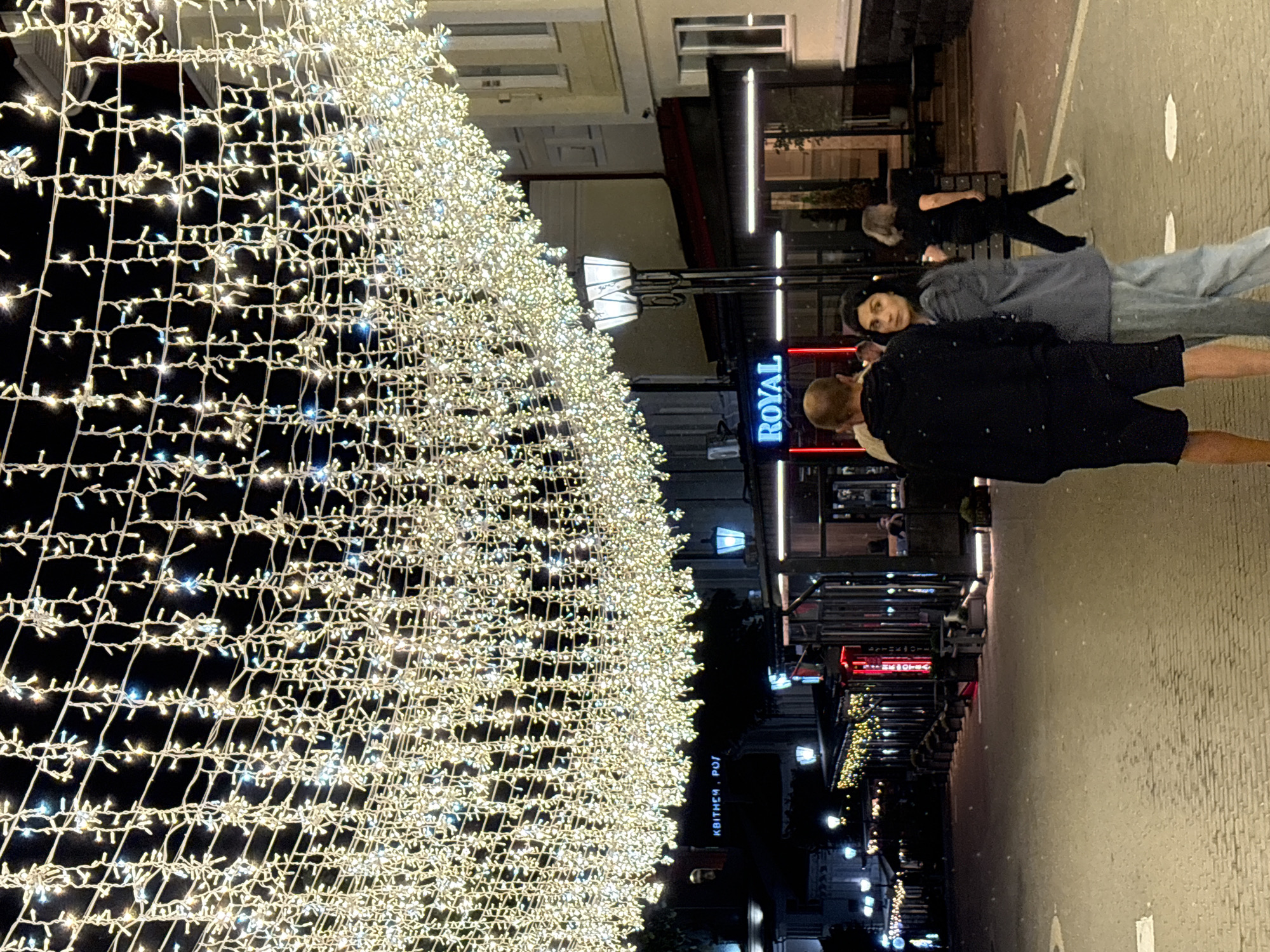 This is a nighttime, eye-level, medium shot of a street scene decorated with an overhead canopy of white string lights. In the foreground, two people, a man and a woman, are walking away from the camera, looking to their right. The man is wearing a black hoodie and shorts, and the woman is wearing a grey blazer and light blue pants. Further down the street, another person, a woman with grey hair, is walking away from the camera towards the right side of the frame. Several businesses with illuminated signs are visible along the sides of the street, including one with a blue neon sign that reads "ROYAL". The street is paved with cobblestones, and there are street lamps and other decorative lights visible. The scene appears to be a lively urban area, possibly a pedestrian street or a marketplace, decorated for an evening event. The location is the street of Sovetskaya in Vitsebsk, Belarus.