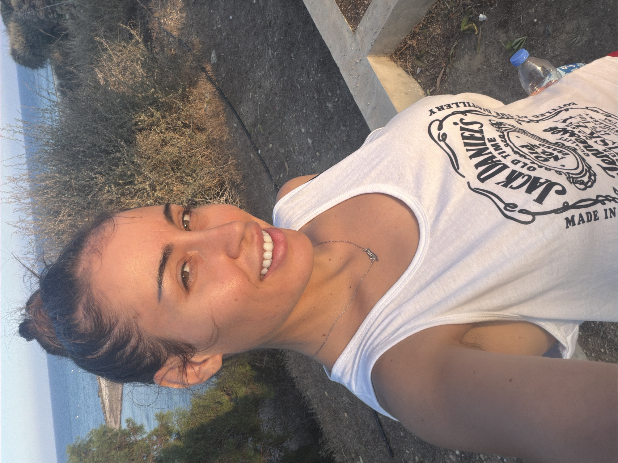 A woman is taking a selfie outdoors in Meneou, Cyprus. The background shows the sea and dry vegetation. The woman is wearing a white tank top with a Jack Daniel's logo. She has her hair pulled up in a bun and is wearing a necklace with the name "Mama". A bottle of water is visible on the ground next to her.