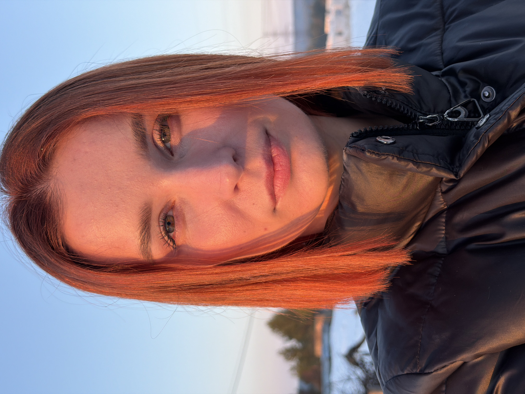 A female individual, appearing to be in her 20s or 30s, is captured in a close-up portrait outdoors in Chaikovsky, Russia. She possesses shoulder-length, straight, reddish-orange hair and fair skin. Direct, low-angle sunlight illuminates the left side of her face (viewer's left) with a warm golden-orange glow, creating a sharp shadow line across the right side. Her eyes are open, revealing light brown or hazel irises, and her lips are naturally pink and slightly parted. She maintains a direct, neutral gaze towards the camera. She is wearing a dark, presumably black, puffer jacket with a visible zipper and snap buttons on the collar. The background features a clear sky, transitioning from a pale blue overhead to a warmer, yellowish hue near the horizon, consistent with either sunrise or sunset. The ground is visibly covered in snow, indicating a winter setting. In the distant background, evergreen trees, a partially obscured wooden structure, and faint utility poles are discernible, suggesting a location on the outskirts of the city.
