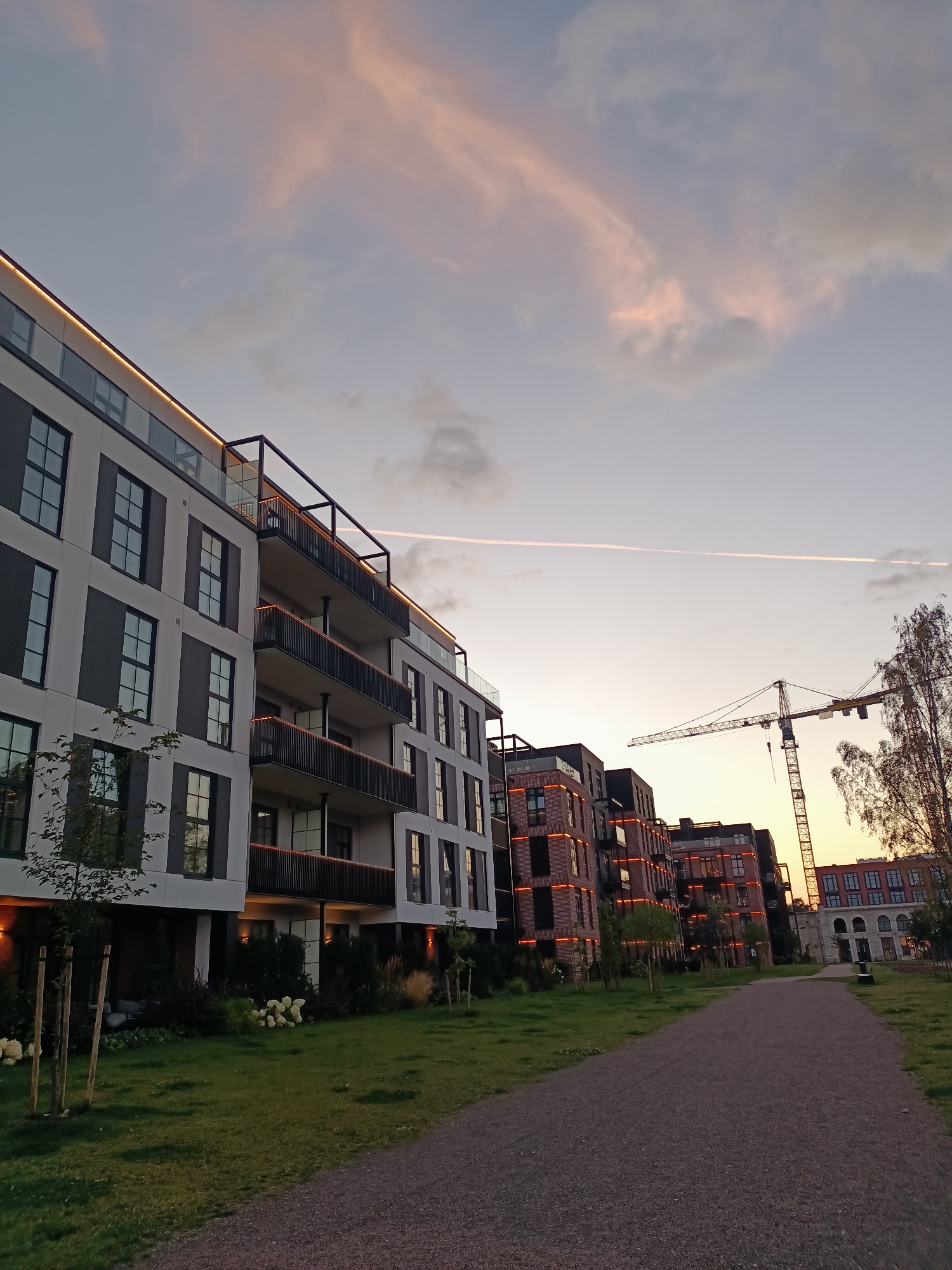 The image depicts a contemporary residential complex under construction in Tallinn, Estonia, likely in a redeveloped urban area such as the Noblessner Quarter. The scene captures a dusk or dawn setting with a pastel-colored sky, tinged with pink and orange hues, indicative of a sunset or sunrise. A faint contrail stretches across the sky, suggesting air traffic.
In the foreground, a gravel path curves gently to the right, flanked by a neatly manicured lawn. Several young trees with thin trunks are planted along the edge of the grass.
Dominating the left side of the frame is a modern apartment building, characterized by its white and dark gray facade, large windows, and multiple balconies with dark railings. Subtle warm lighting illuminates the balconies and the upper edge of the building, adding a touch of ambiance.
Further to the right, a row of brick buildings with illuminated orange accents stands out, hinting at the ongoing development. A large construction crane looms over these buildings, a clear indicator of ongoing construction activities. To the far right, a more traditional, older stone building with arched windows is partially visible, suggesting a blend of old and new architecture in the area. The overall atmosphere is one of urban renewal and modern living. No people are visible in the image.