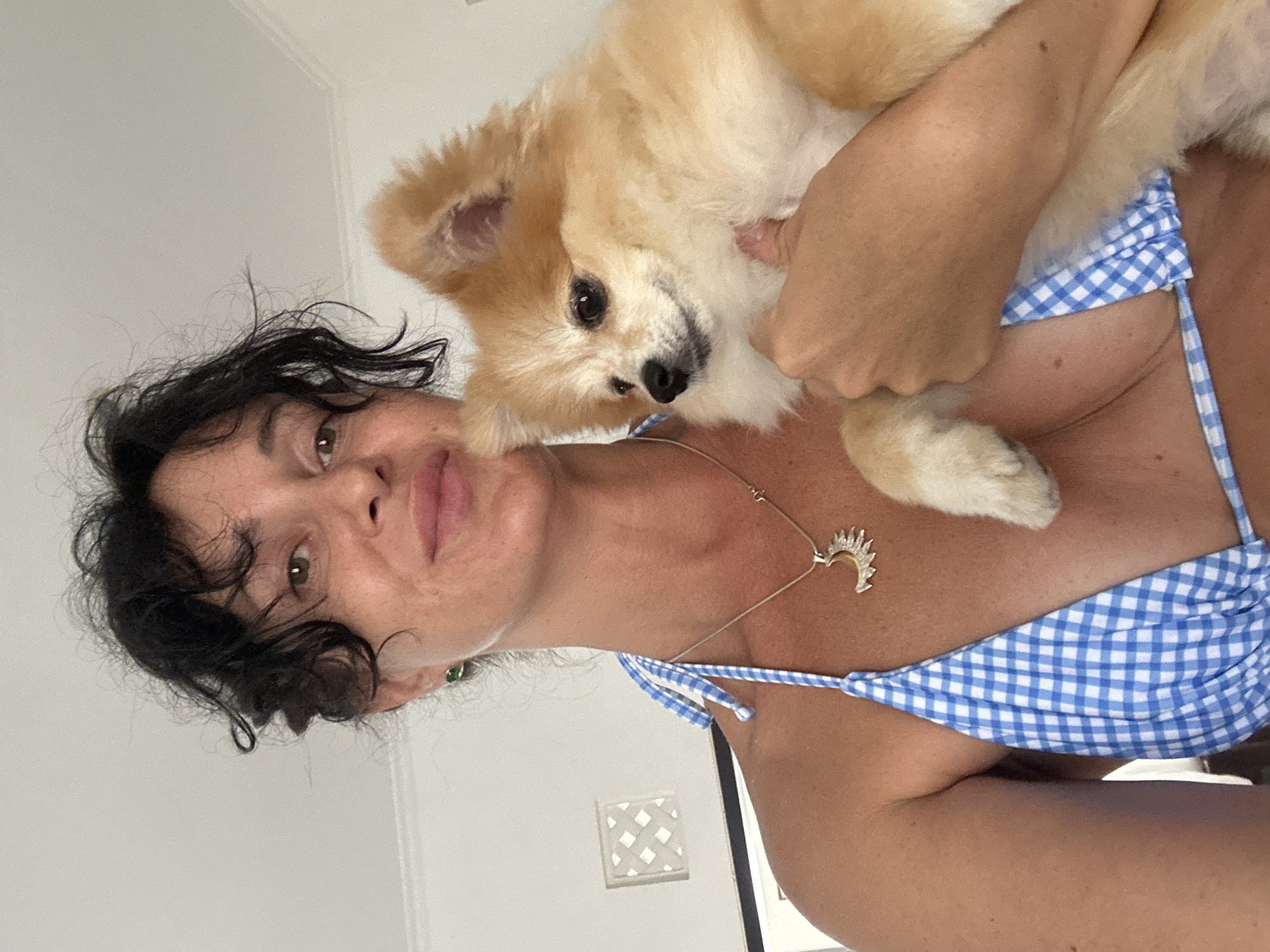 In a close-up, self-taken photograph, a woman with dark, curly hair and light brown eyes is holding a small, fluffy Pomeranian dog. She is wearing a blue and white checkered bikini top and a silver necklace with a sun-shaped pendant. The woman is looking slightly to the right, with a subtle smile. The dog, with light brown and white fur, is looking to the left. They are indoors, with a plain white wall and a decorative square panel visible in the background. The lighting suggests an indoor setting, possibly during the daytime. The overall impression is of a casual, intimate moment between the woman and her pet, with the location being Kuta Selatan, Indonesia.