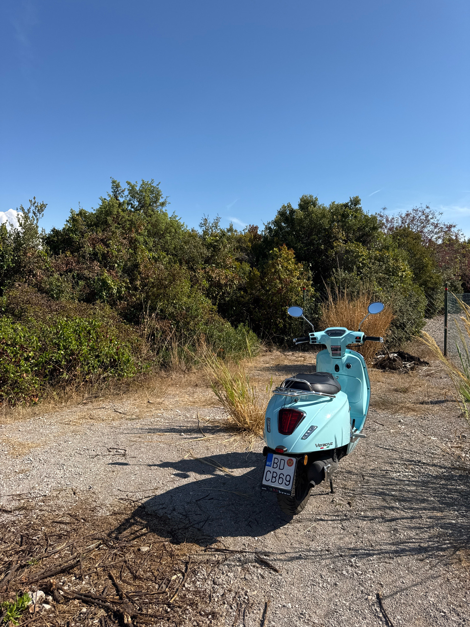 A light blue scooter is parked on a gravel path in a rural area. The scooter has a license plate with the text "BD CB69". The background consists of dense green foliage and trees under a clear blue sky. The path appears to be unpaved and is surrounded by dry grass and scattered branches. The image captures a sunny day with clear visibility. The location is identified as Dubraljevina, Montenegro.