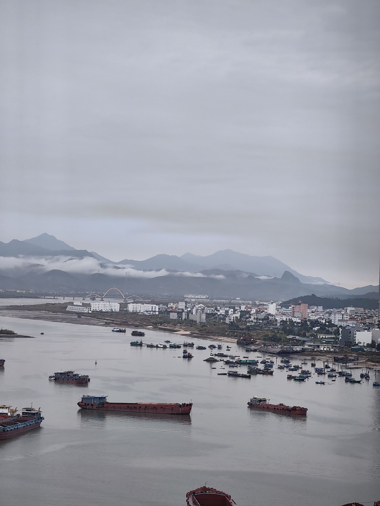 An aerial shot shows a river scene in Hong Gai Ward, Vietnam, on a cloudy day. Mountains are visible in the background. Numerous boats are scattered across the river, varying in size and purpose, including larger barges and smaller fishing vessels. Buildings and infrastructure are visible along the river's edge, including bridges.