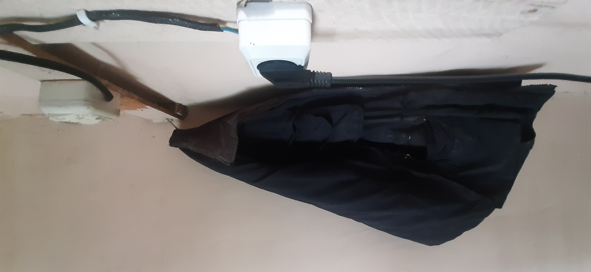 A black jacket hangs from a wooden peg on a light-colored wall. Two electrical outlets are visible on the wall. One outlet, located above and to the left of the jacket, appears to have a black cable plugged into it, with a portion of the cable secured by a white clip. The second outlet, located to the right of the jacket, has a black plug inserted into it. A thick black cable extends from this plug downwards. The wall itself appears somewhat worn and textured. The image is taken from a slightly low angle, looking up towards the hanging jacket and outlets. The scene is indoors. The location is Yessentukskaya, Russia.