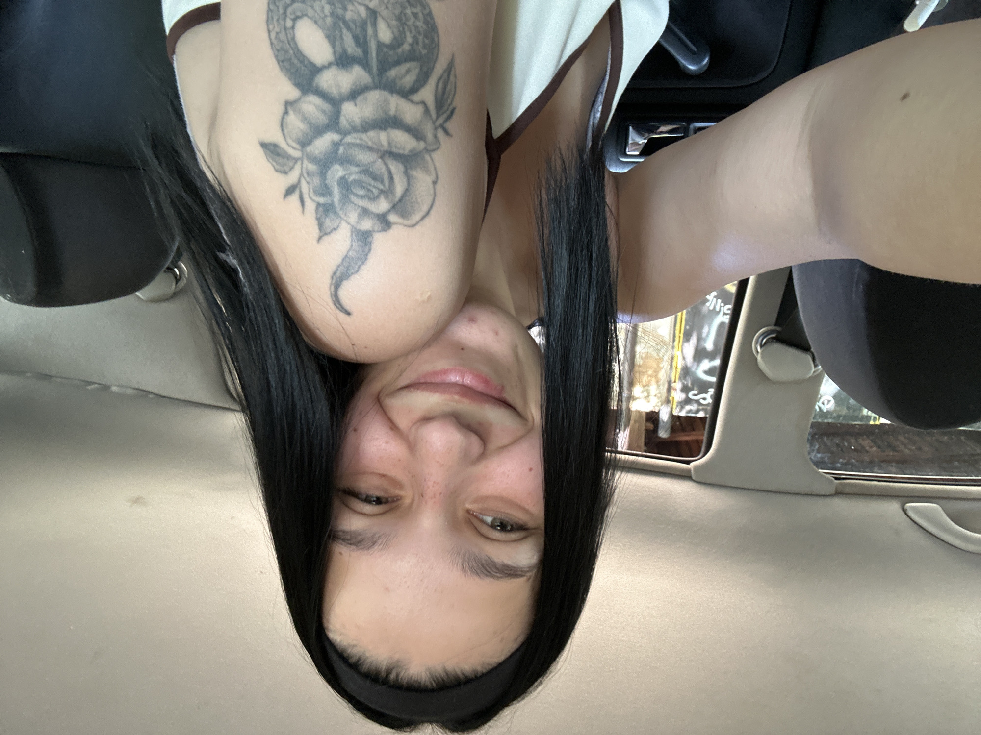 A young adult female is depicted taking a selfie inside a vehicle. She has long, straight, black hair, worn with a black headband. Her face is smiling towards the camera, revealing slight freckles and a small blemish on her chin. She is wearing a light-colored camisole with thin brown shoulder straps. A detailed black and white tattoo featuring a rose intertwined with a snake is prominently visible on her left upper arm.
The interior of the vehicle shows a light beige, textured fabric headliner, with a matching grab handle and a B-pillar trim piece. Dark-colored car seats are visible on both the left and right sides of the frame, with the headrest of the passenger seat partially visible on the right. A portion of the car's door panel, featuring black plastic and metallic trim elements, is also visible on the right.
Through the side window on the right, an outdoor urban environment can be seen. The background consists of weathered walls or structures with white graffiti or markings, including recognizable letters such as "S.N." and "v". The lighting indicates a daytime setting. The scene is located in a vehicle in Sofia, Bulgaria.