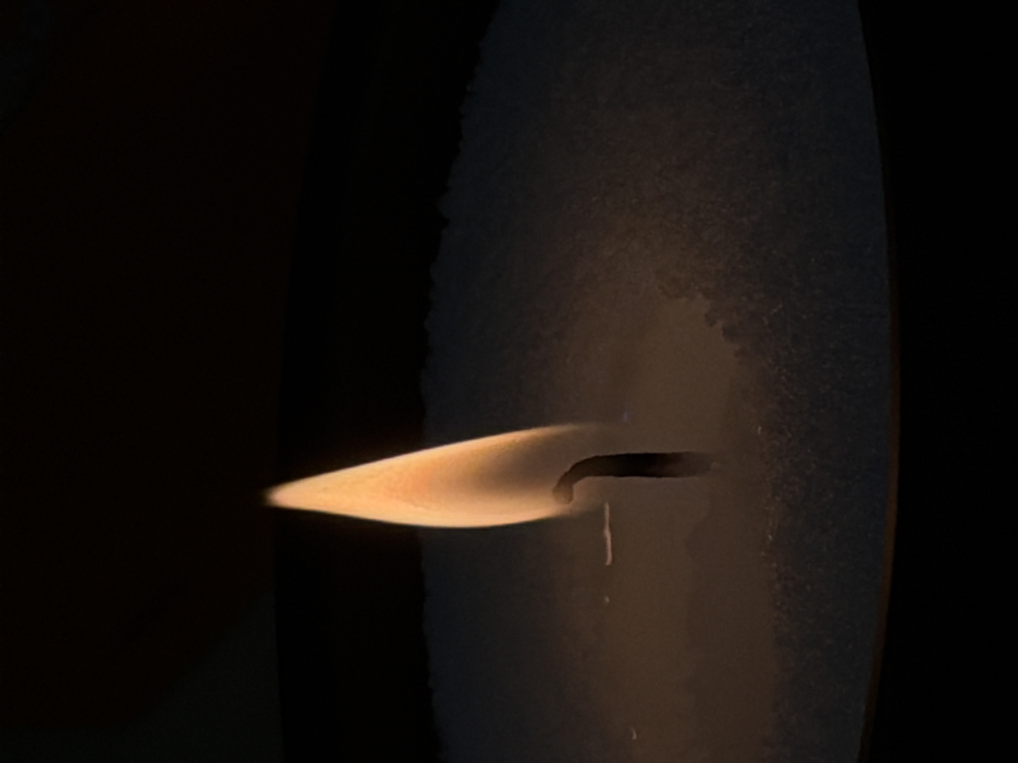 This is a close-up, low-angle shot of a lit candle with a dark background. The flame is a bright orange and yellow, tapering to a point at the top. The wick is visible as a dark, thin line extending down from the flame into a powdery substance that appears to be the candle's wax. The wax is textured and forms a shallow, circular base around the wick. The lighting is dim, with the flame being the primary light source, casting a warm glow on the immediate surroundings. The overall impression is one of simplicity and quietude. There are no people, animals, or discernible elements of Ashkelon, Israel, present in the image.