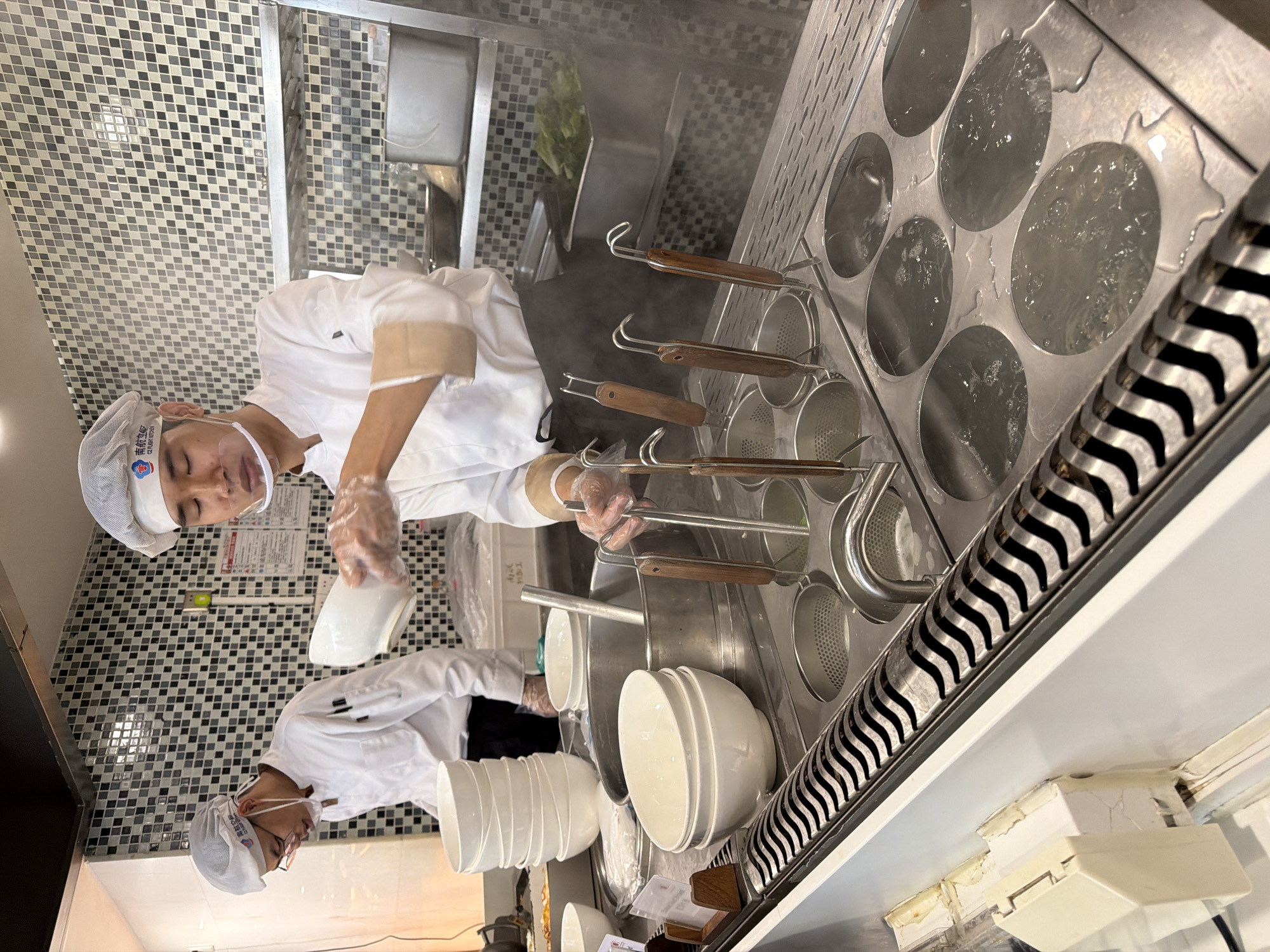 The image depicts an indoor scene within a commercial kitchen, likely located in Baiyun District, China. A chef, dressed in a white chef jacket, black apron, white hairnet, clear face shield, and plastic gloves, is actively engaged in food preparation. The chef is holding a white ceramic bowl, appearing to be in the process of serving or preparing a dish, likely noodles, given the surrounding equipment.
The central part of the scene features a large stainless steel noodle cooker or boiler with multiple circular openings. Several of these openings contain boiling water, from which steam is visibly rising. Other openings hold metal noodle strainers with wooden handles, indicating a noodle-making operation. To the right of the cooker, there's a distinctive metal grate with a wavy pattern. Stacks of clean white ceramic bowls are arranged on the stainless steel counter, ready for use.
The background wall is tiled with small square tiles in a mosaic pattern of black, grey, and white, typical of a professional kitchen. Stainless steel sinks and surfaces are also visible. A reflection in the lower part of the image shows another chef (or the reflection of the same chef) wearing a similar uniform and glasses, reinforcing the impression of a busy food preparation area.
A significant detail is the visible text on the main chef's white hairnet, which reads "南航食品" (Nán Háng Shí Pǐn). This translates to "China Southern Airlines Food," suggesting the establishment is a catering service or restaurant associated with China Southern Airlines. The overall scene conveys a focused and organized environment for food preparation.