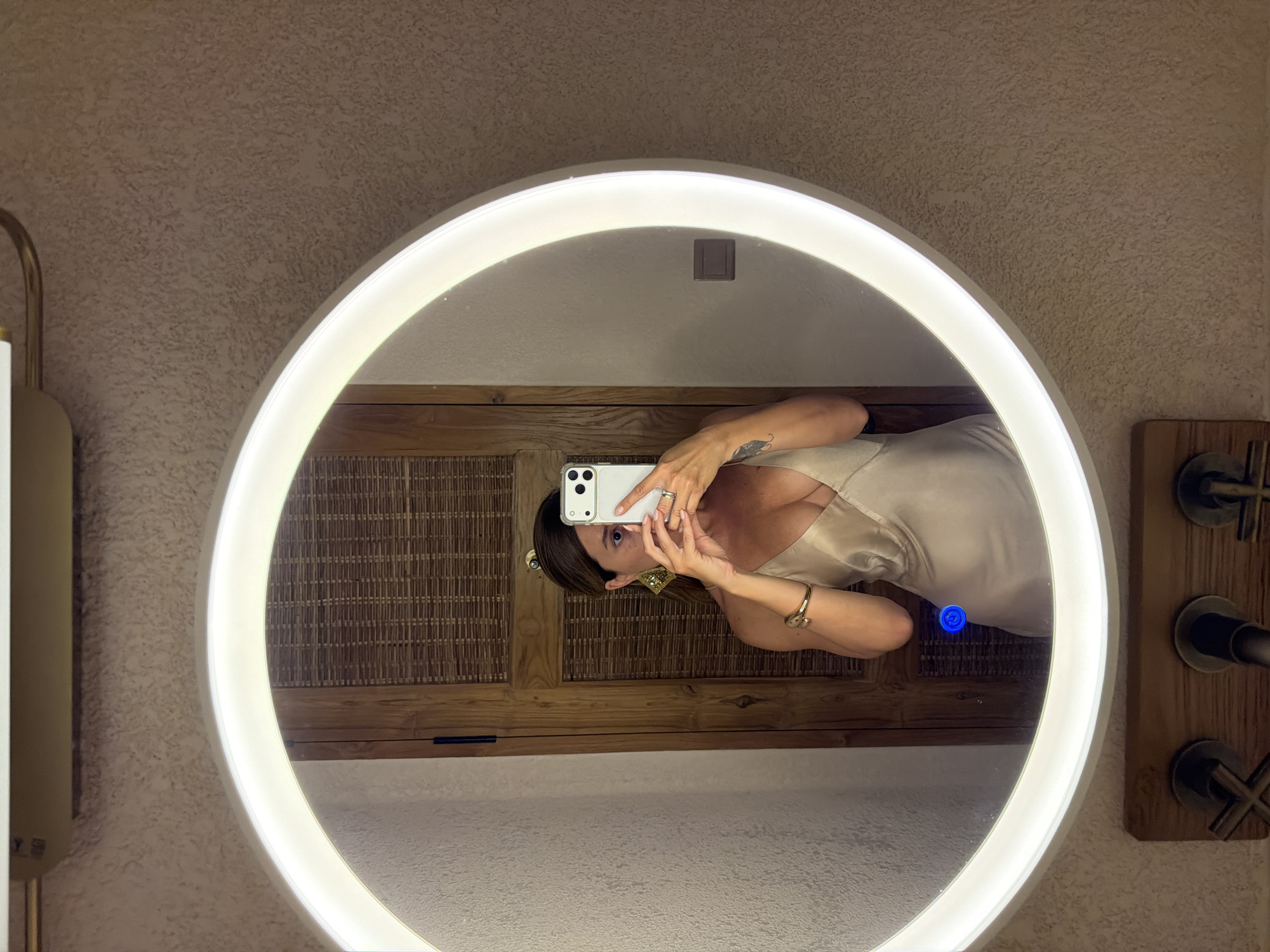 The image is a mirror selfie taken in a bathroom. The subject is a person, reflected in a large, circular, backlit mirror. The person is holding a white smartphone in their right hand, using it to take the picture. They are wearing a light-colored satin dress with a low neckline and jewelry, including rings and a bracelet. The background reflected in the mirror shows a wooden cabinet or wall paneling with a woven texture, and a wall with textured plaster. A blue light is visible in the reflection, possibly from a button on the mirror or nearby. To the right of the mirror, part of a wooden fixture with dark metal knobs is visible, suggesting bathroom faucets. The overall lighting is warm and ambient, consistent with indoor lighting. The setting appears to be a modern bathroom with natural materials. The photo is taken from a low angle, looking up at the mirror. The time of day and weather are not discernible. There is no visible text in the image. The location context is Mengwi, Indonesia.