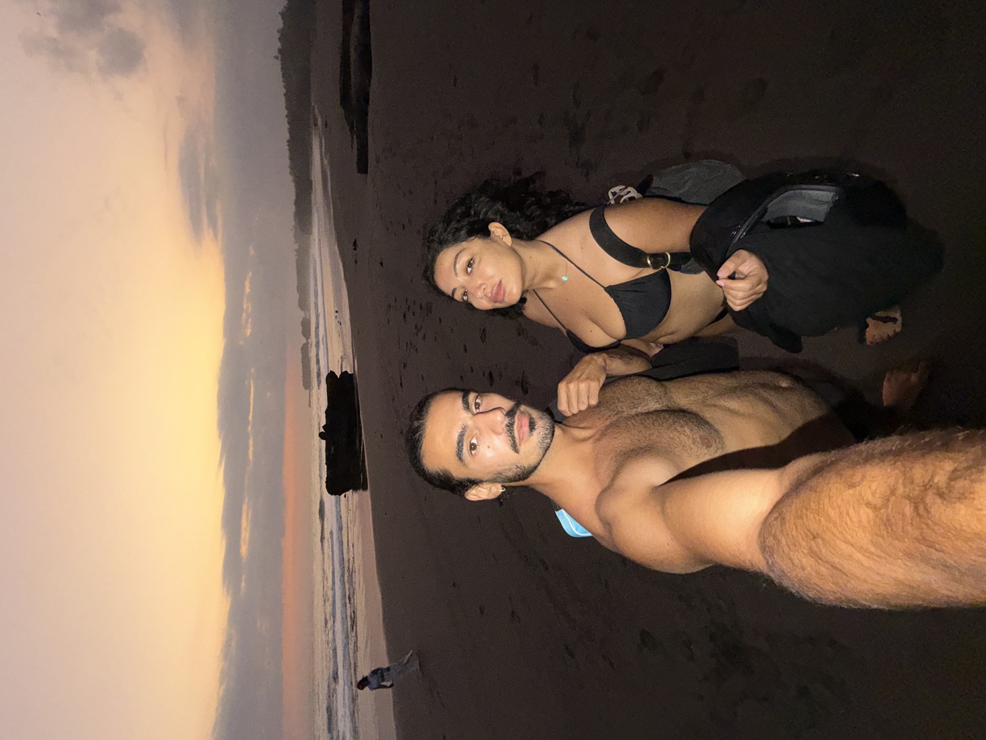 The image depicts a beach scene in Selemadeg Timur, Indonesia, during twilight hours. In the immediate foreground, two individuals are captured in what appears to be a selfie.
The man, positioned on the left, is bare-chested, exhibiting dark hair pulled back, a beard, and a mustache. He wears a small earring in his left ear, and a light blue strap is visible over his left shoulder. His right arm is extended towards the camera, suggesting he is holding the device.
To his right, the woman has dark, curly hair and is wearing a black bikini top with a light-colored necklace featuring a small turquoise pendant. A black strap, likely from a bag, is visible on her left shoulder, and she holds a dark jacket or garment in her right hand. Both individuals are looking directly at the camera.
The background showcases a dark sand beach with gentle waves breaking on the shore. A large, dark rock formation is prominently located in the mid-ground, near the water. Further to the left in the distance, a solitary figure dressed in light-colored clothing stands at the water's edge, facing away from the camera towards the ocean. The sky above displays a gradient of warm orange and yellow hues near the horizon, transitioning into pink and purple tones, with several scattered dark clouds. A dark, vegetated coastline is discernible along the far right horizon. The overall lighting is dim, indicating low natural light conditions, with the foreground subjects appearing distinctly illuminated by a direct light source, likely originating from the camera.