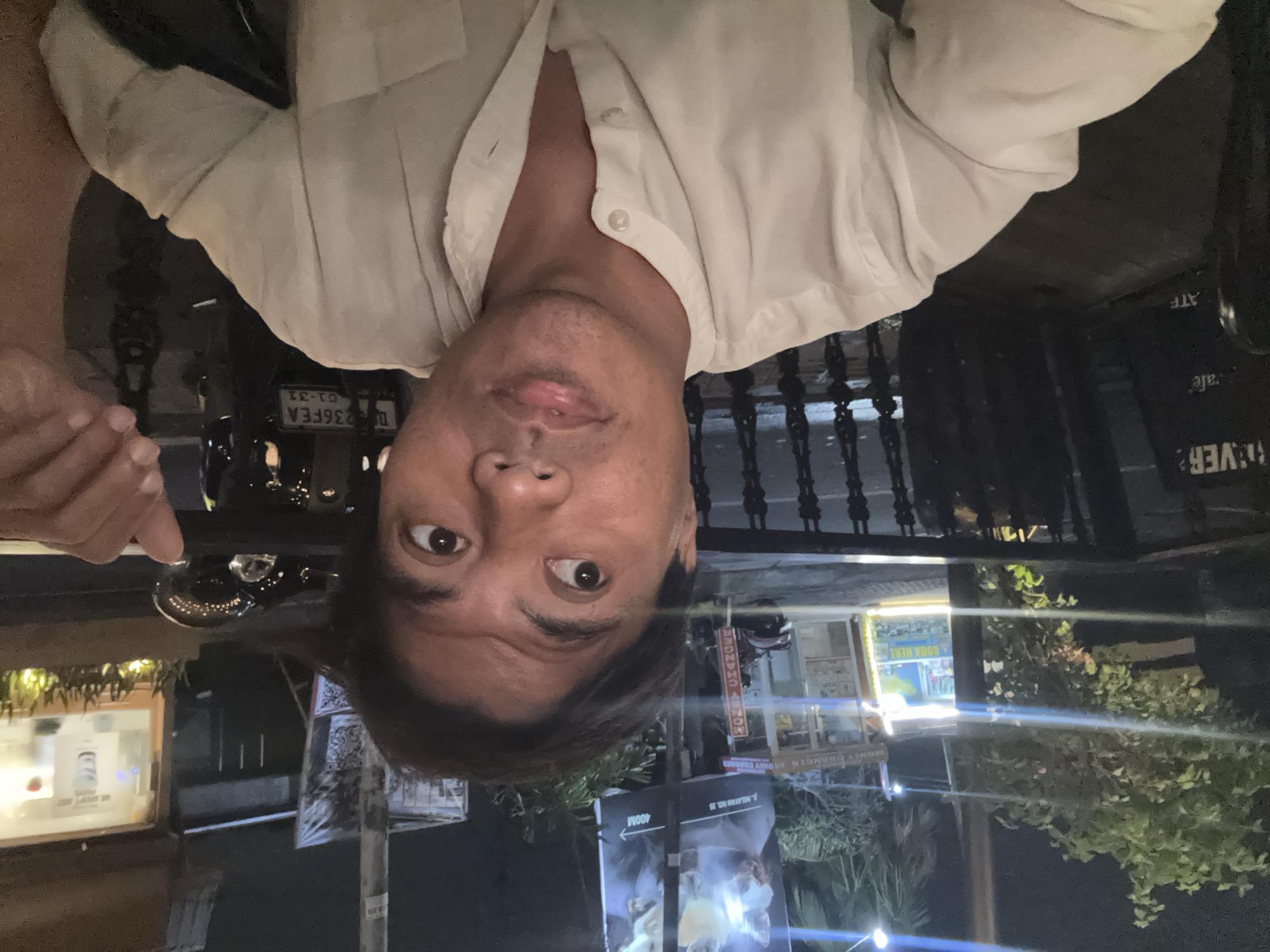A selfie taken by a man in his 30s or 40s at night in North Kuta, Indonesia. The man is wearing a light-colored, collared shirt. He is looking off to the right side of the frame with a slightly open mouth. The background shows a street scene with shops, a billboard, and parked motorcycles. The lighting is dim, typical of nighttime street lighting, with some bright highlights from neon signs and vehicle lights. The overall atmosphere suggests an urban setting at night, possibly a tourist or entertainment district.