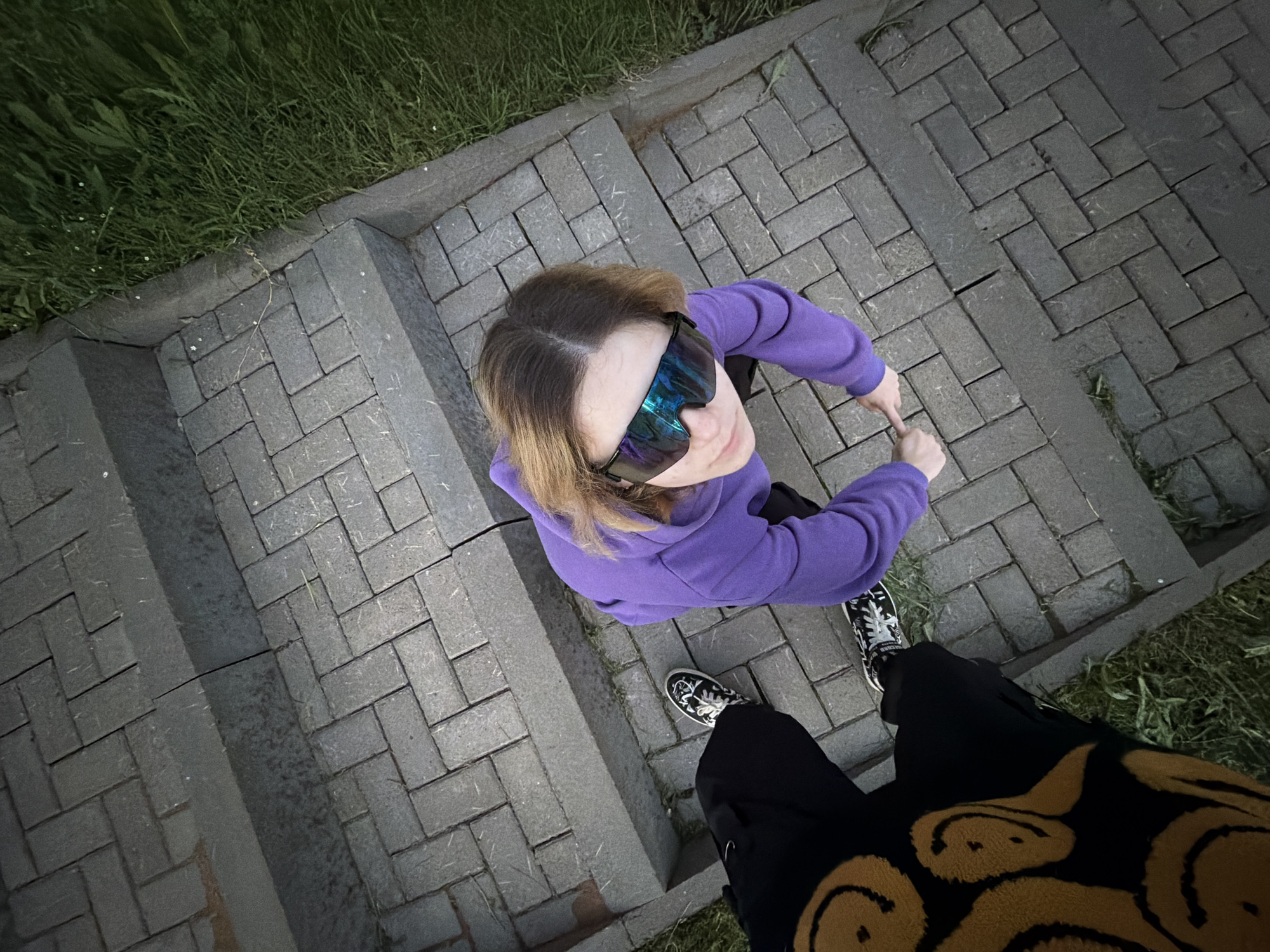 An overhead shot captures two individuals standing on a brick pathway bordered by grass. The person in the foreground, wearing black pants with a smiley face pattern, is facing away from the camera. To their right, another person, seen from the chest up, is looking upwards. This individual is wearing a purple hooded sweatshirt and reflective goggles. Both individuals are wearing sneakers. The scene appears to be outdoors, possibly a park or a paved walkway, with hints of greenery suggesting a natural environment. The lighting suggests it could be late afternoon or early evening. There are no visible text elements in the image.