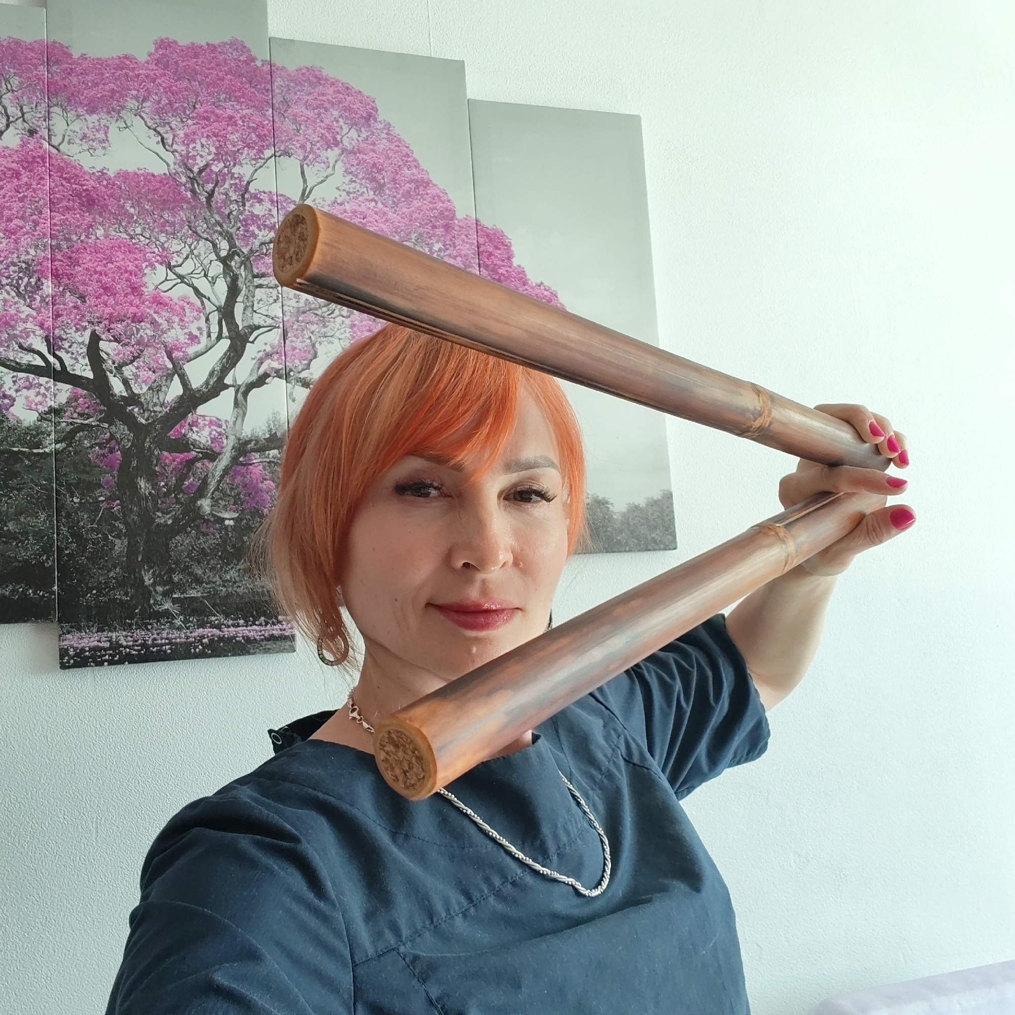 A fair-skinned woman with short, reddish-orange hair is holding two wooden bamboo sticks in front of her face. She is wearing a dark blue shirt and a silver necklace. Her fingernails are painted bright pink. Behind her is a piece of artwork depicting a tree with pink blossoms; one panel of the artwork is in grayscale. The photograph was taken in an interior space with white walls. While I am unable to determine from this image that the location is in Chaikovsky, Russia.