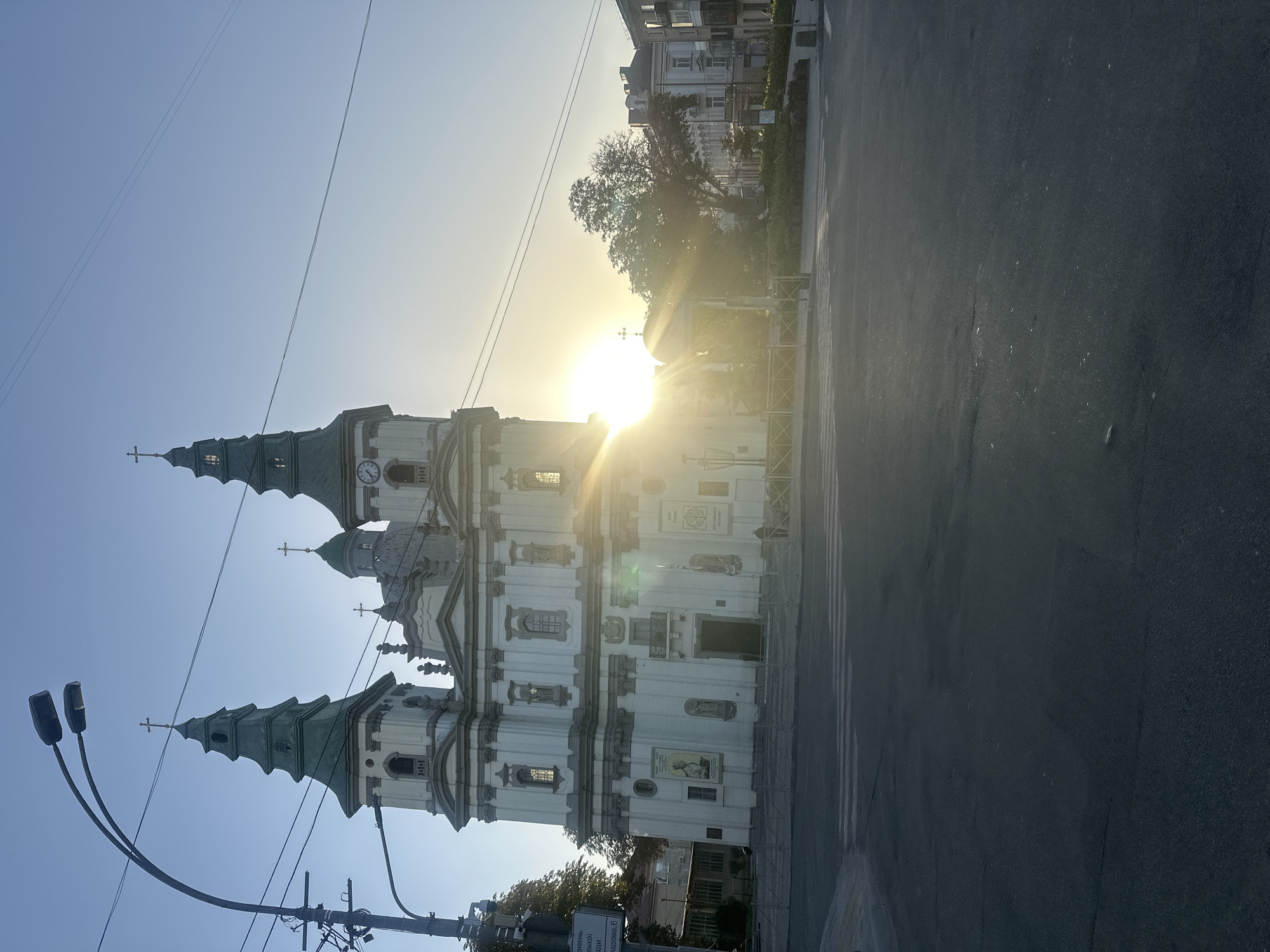 The image shows a large, white, ornate church with two green-topped spires. The sun is setting and is directly behind the church, creating a bright lens flare. The church has many windows and decorative elements, including statues and a clock face on one of the spires. There is a street in front of the church with a pedestrian crossing. A person can be seen standing on the sidewalk in front of the church. The sky is clear and blue, with a few power lines visible. Trees and other buildings are visible to the right of the church. The image appears to have been taken from a low angle, looking up at the church. The location is the Ternopil Theological Seminary of the Order of Saint Basil the Great in Ternopil, Ukraine.