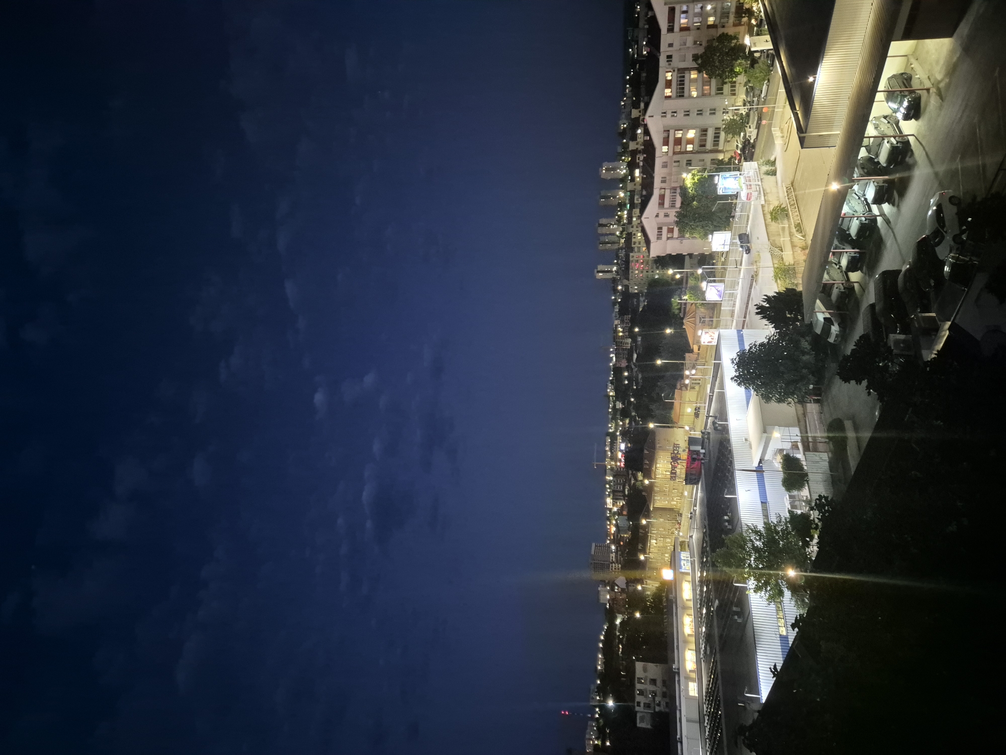 A nighttime aerial view of an urban landscape with a dark blue sky overhead, featuring scattered clouds. In the foreground, a dark, leafy foreground element is visible, obscuring part of the scene. Below, a large commercial building with a white roof and illuminated interior spaces dominates the lower section of the image. To the right of this building is a covered parking area where several vehicles, including vans and cars, are parked under artificial lighting. Further into the distance, a sprawling cityscape unfolds with numerous buildings of varying heights, many of them residential apartments with illuminated windows. Streetlights dot the landscape, creating a network of light across the urban area. In the far distance, taller buildings and a crane are faintly visible against the twilight sky. The overall impression is of a moderately sized city at dusk or early night.