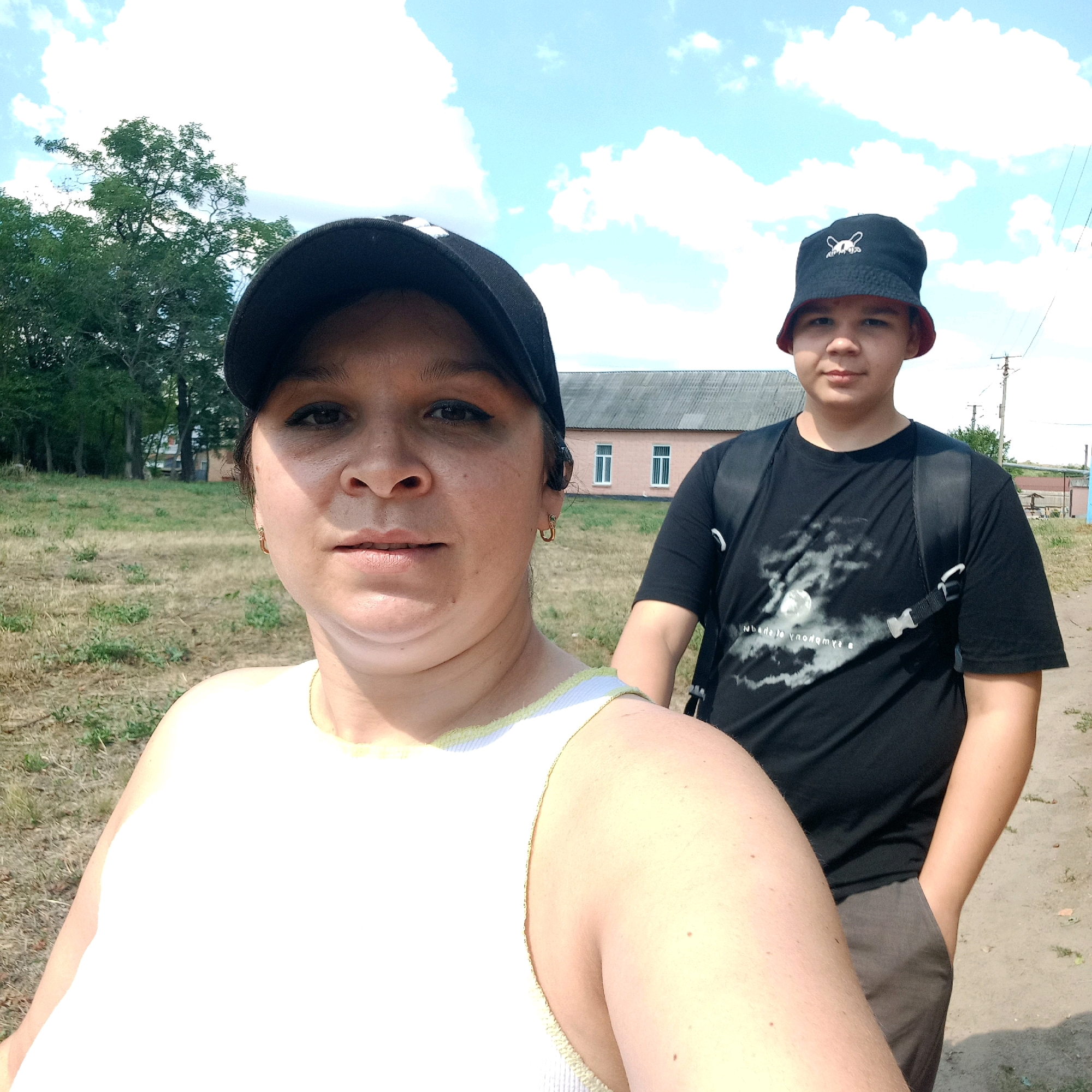 In the city of Klyntsi, Ukraine, on a sunny day with scattered clouds, a woman and a boy are posing for a selfie outdoors. The woman, in the foreground, wears a black baseball cap and a white tank top. To her right, the boy sports a dark bucket hat with a white cartoon design and a black t-shirt. He has a backpack on. They are standing on a dirt path, with dry grass and trees visible behind them. In the background, a long, single-story building with a gray roof and two rows of windows can be seen. A utility pole stands to the right of the building. The overall scene suggests a casual outdoor activity or outing in a semi-rural area.